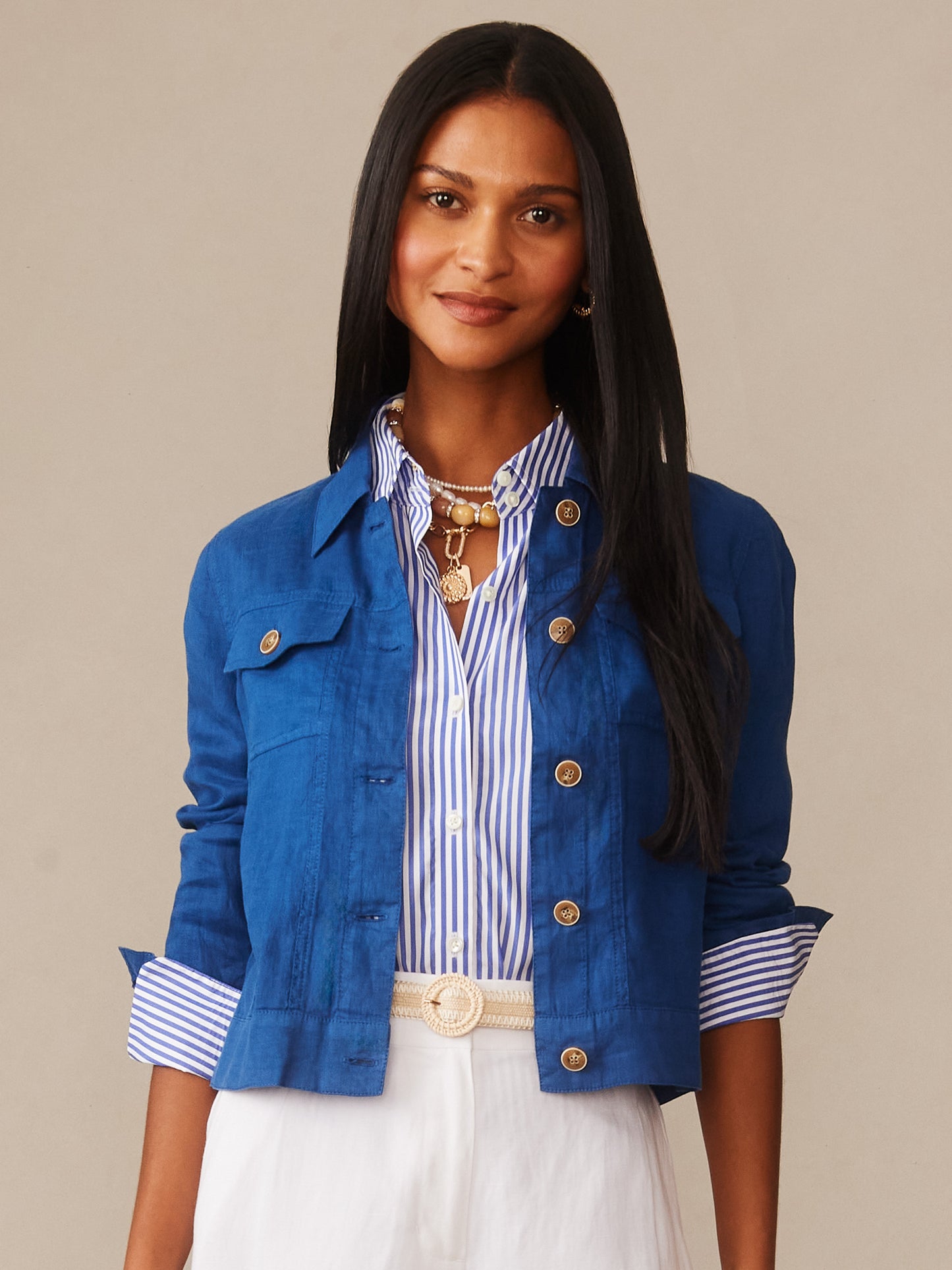 A woman wearing a vibrant blue Halo Linen Jacket over a striped shirt, styled with white pants and a beige belt, showcasing J.McLaughlin’s effortlessly stylish, enduring designs and innovative fabrics.
