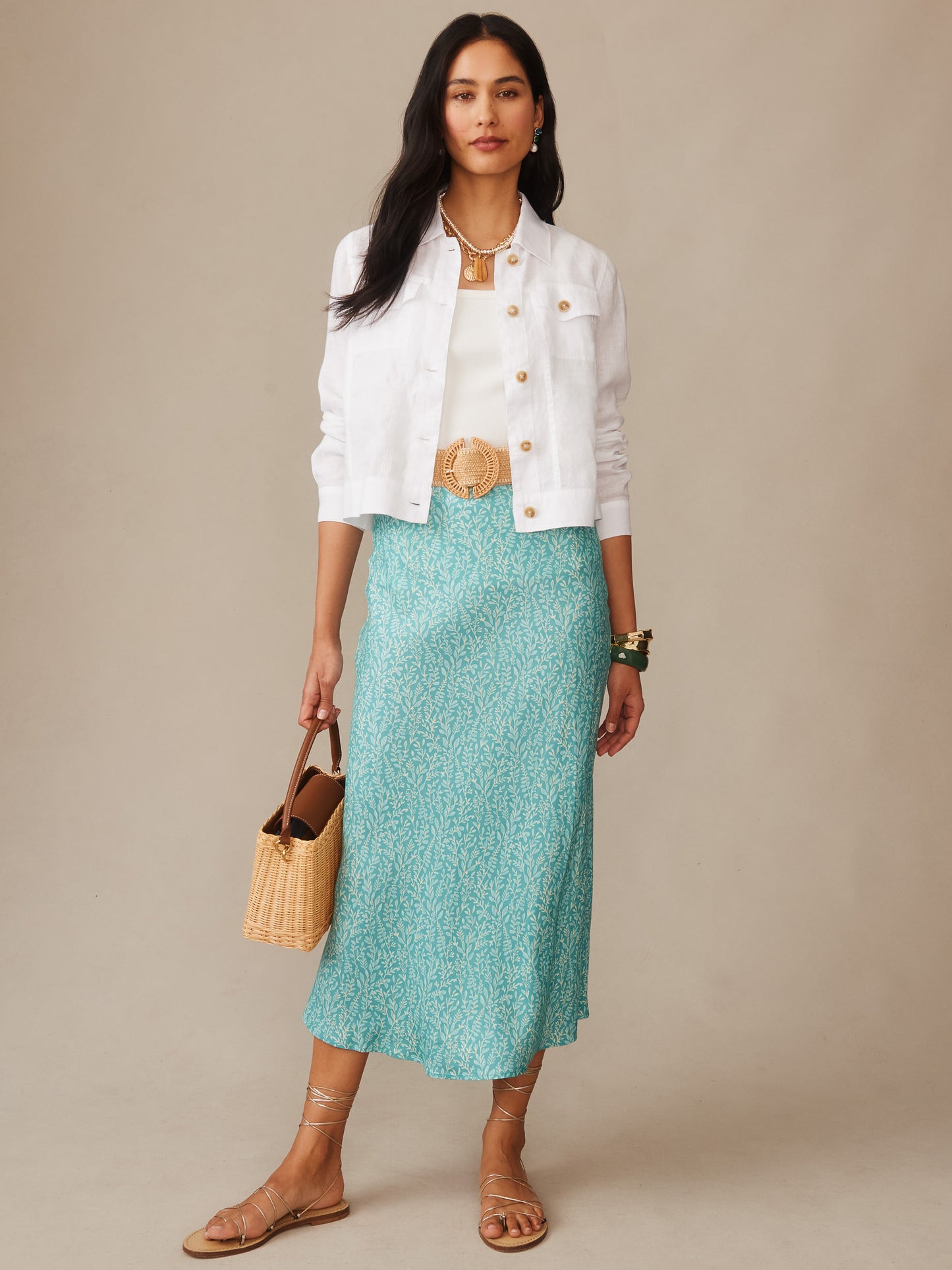 Woman wearing a Halo Linen Jacket by J.McLaughlin in white over a white top and teal patterned skirt, accessorized with layered gold jewelry, a woven belt, straw handbag, and sandals, showcasing effortlessly stylish, enduring designs with innovative fabrics, a destination for defining style. Life, Well-Lived.