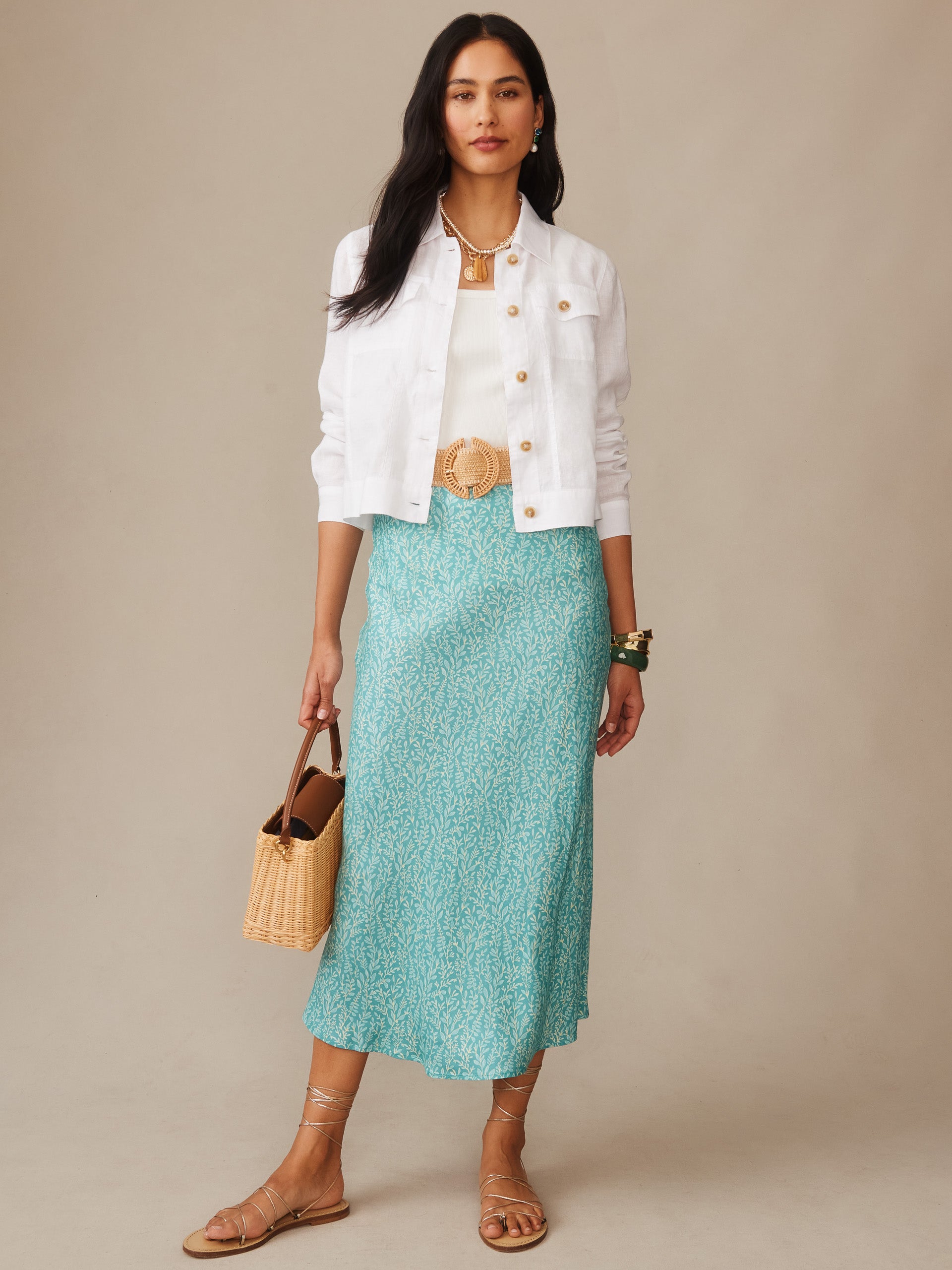Woman wearing a Halo Linen Jacket by J.McLaughlin in white over a white top and teal patterned skirt, accessorized with layered gold jewelry, a woven belt, straw handbag, and sandals, showcasing effortlessly stylish, enduring designs with innovative fabrics, a destination for defining style. Life, Well-Lived.