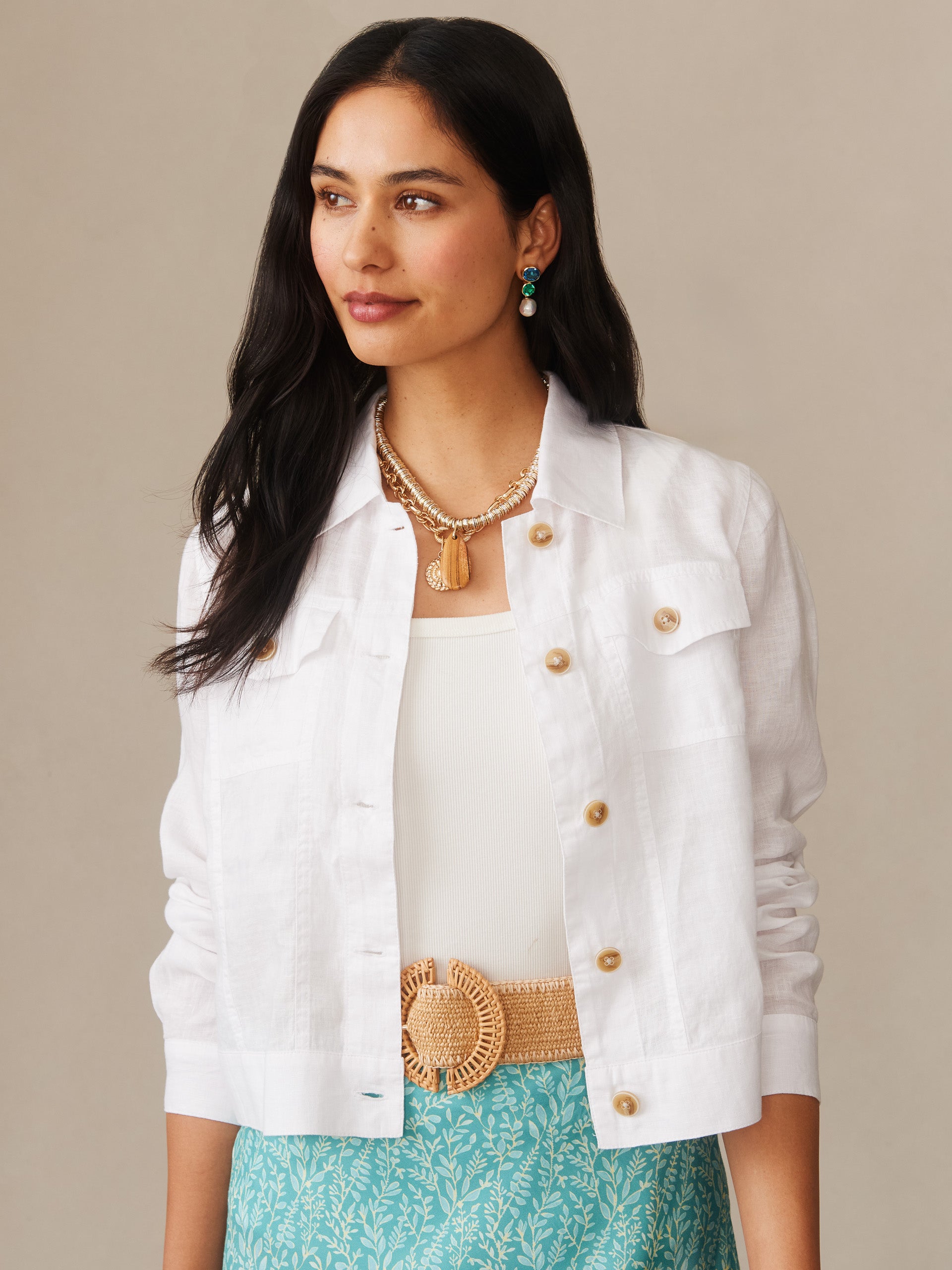 Woman wearing a crisp white Halo Linen Jacket by J.McLaughlin, styled with a woven belt, layered gold necklaces, and vibrant patterned teal skirt, showcasing effortlessly stylish, enduring designs with innovative fabrics for a life well-lived.