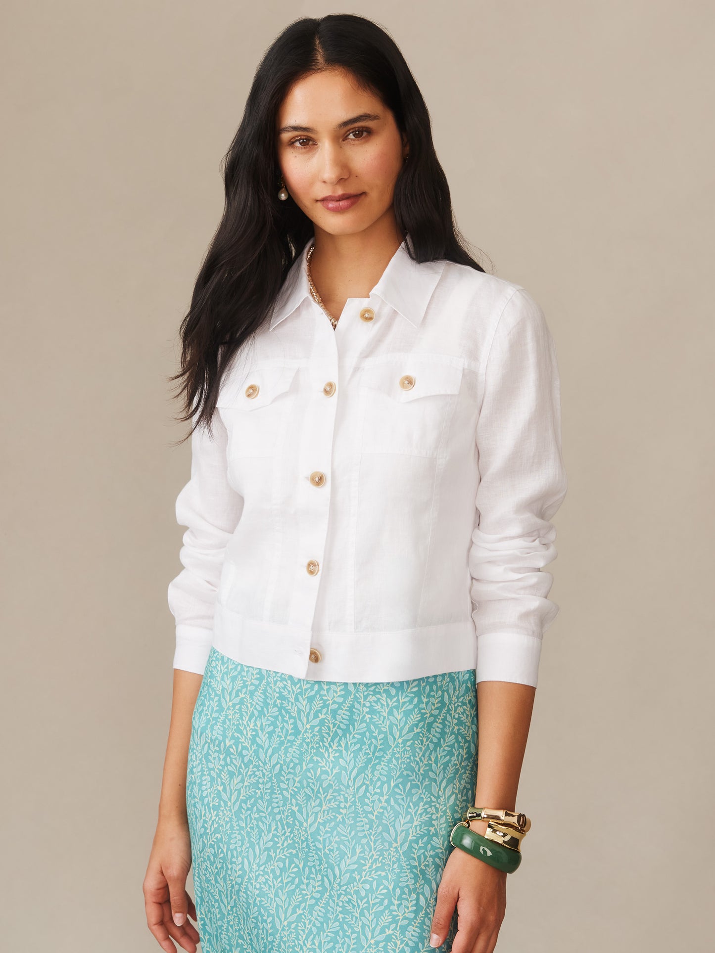 A woman wearing the Halo Linen Jacket in white by J.McLaughlin, paired with a patterned turquoise skirt and stylish bracelets, showcasing effortlessly stylish, enduring designs and innovative fabrics for a life well-lived.