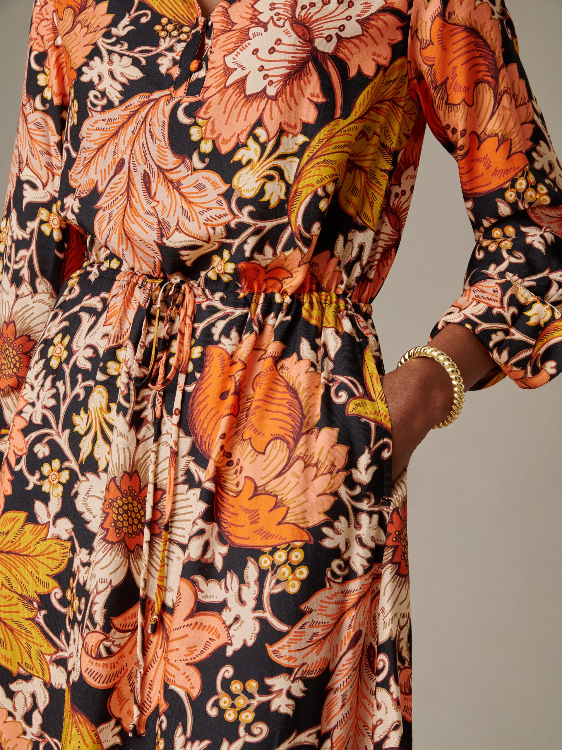 Close-up of a woman wearing the Hannah Silk Blend Dress in City Garden by J.McLaughlin, featuring an elegant floral pattern with warm orange, yellow, and beige tones, and a drawstring waist detail.