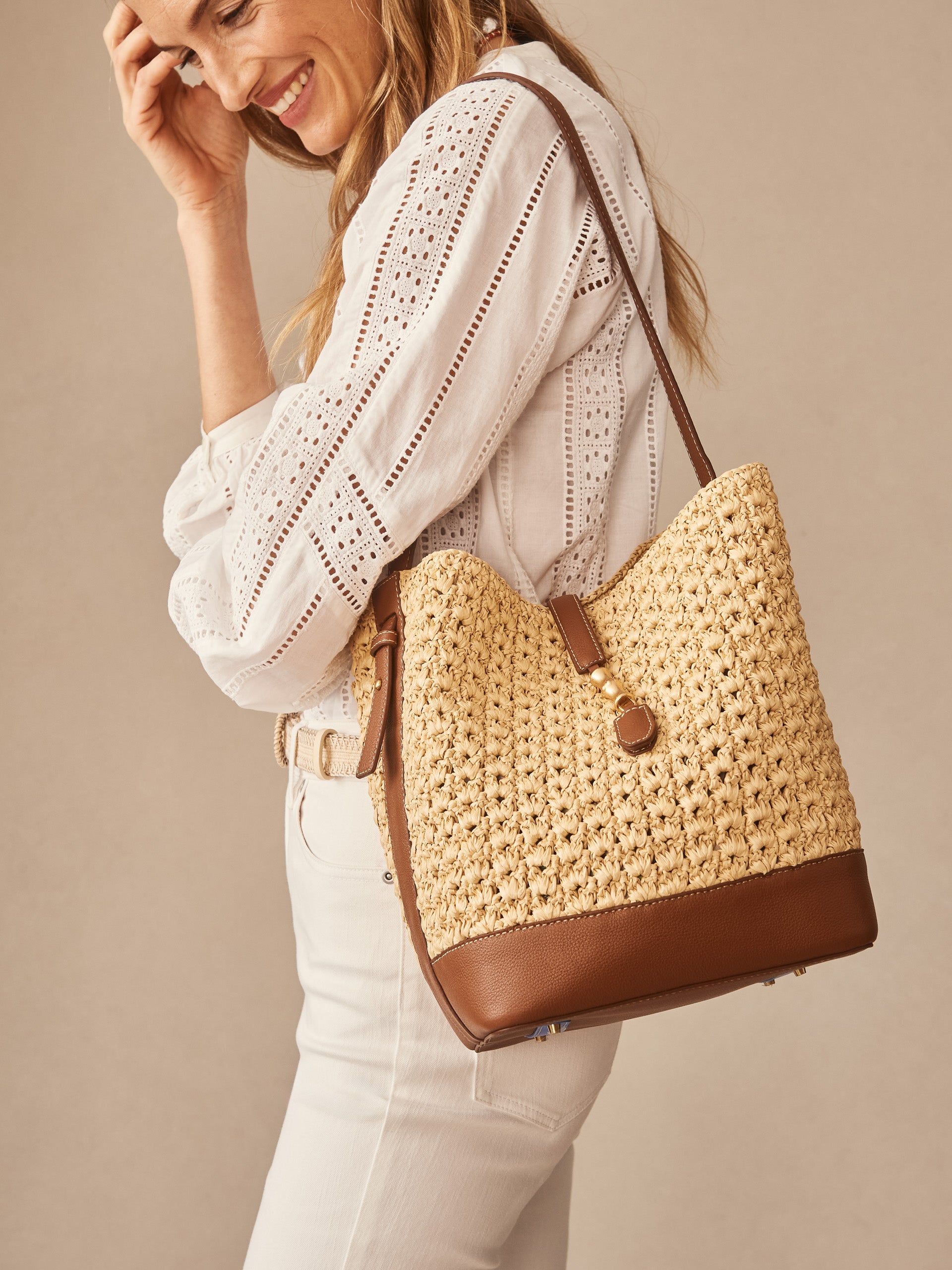 Woman wearing a white eyelet blouse and light pants, carrying a Crawford Raffia Handbag by J.McLaughlin, showcasing effortlessly stylish, enduring designs and a legacy of timeless fashion.
