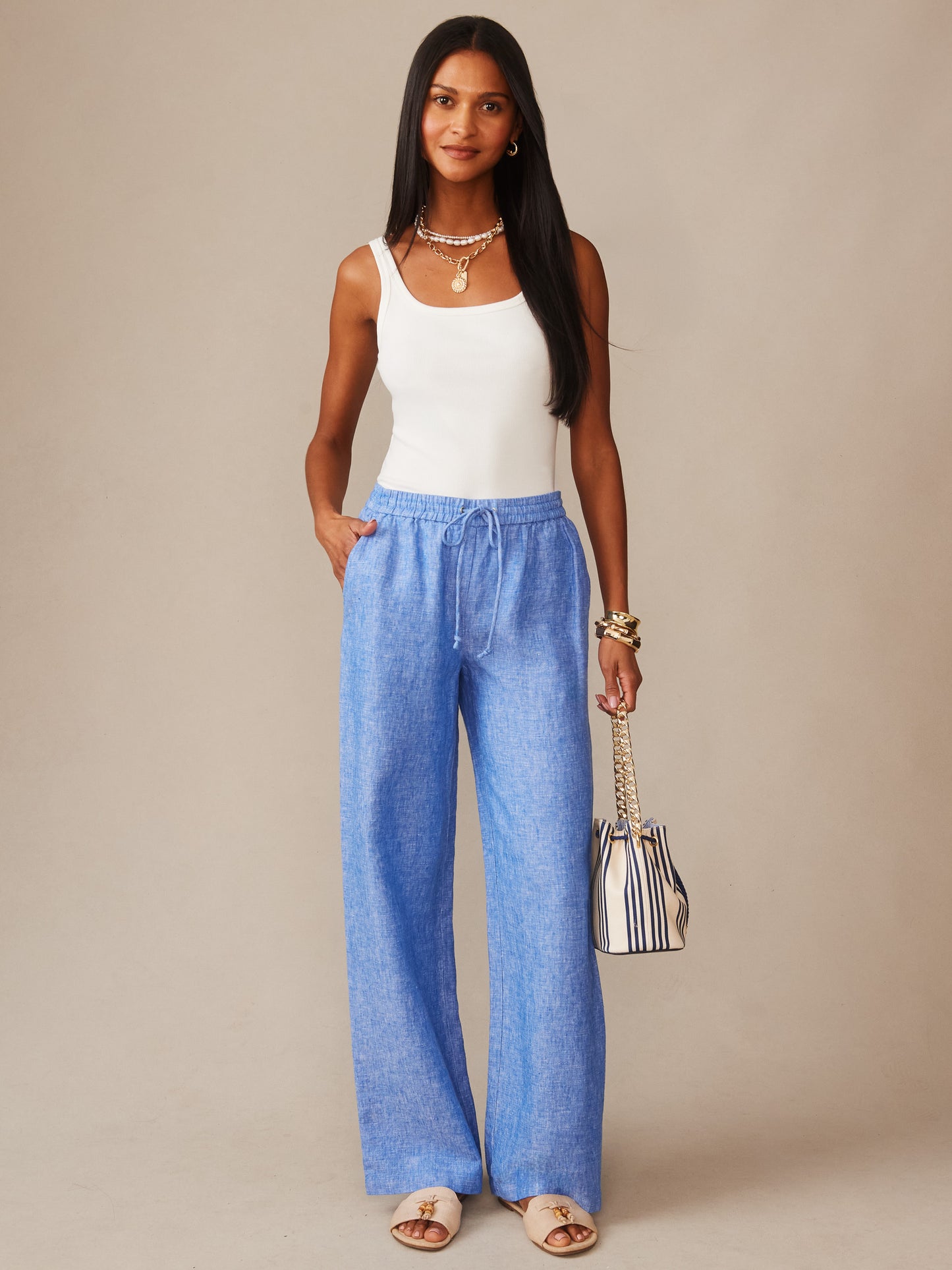 Woman wearing Irene Linen Pants in a soft blue shade by J.McLaughlin paired with a white tank top, accessorized with layered necklaces, bracelets, and a striped handbag. Effortlessly stylish with enduring designs and a legacy of timeless fashion, a destination for defining style. Life, Well-Lived.