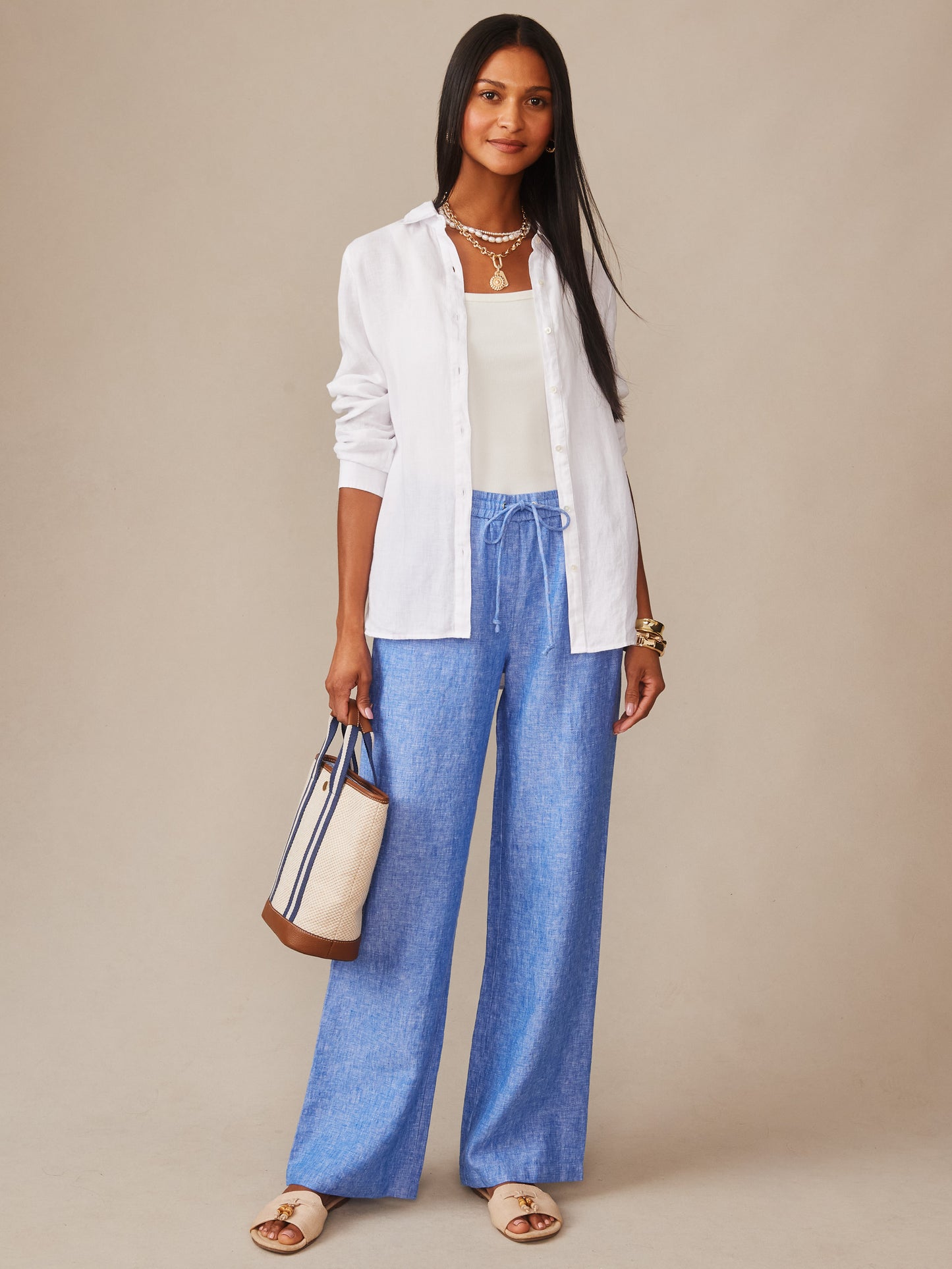A stylish woman wearing J.McLaughlin's Irene Linen Pants in a relaxed fit paired with a white shirt and tank, carrying a chic tote, embodying effortlessly stylish, enduring designs.