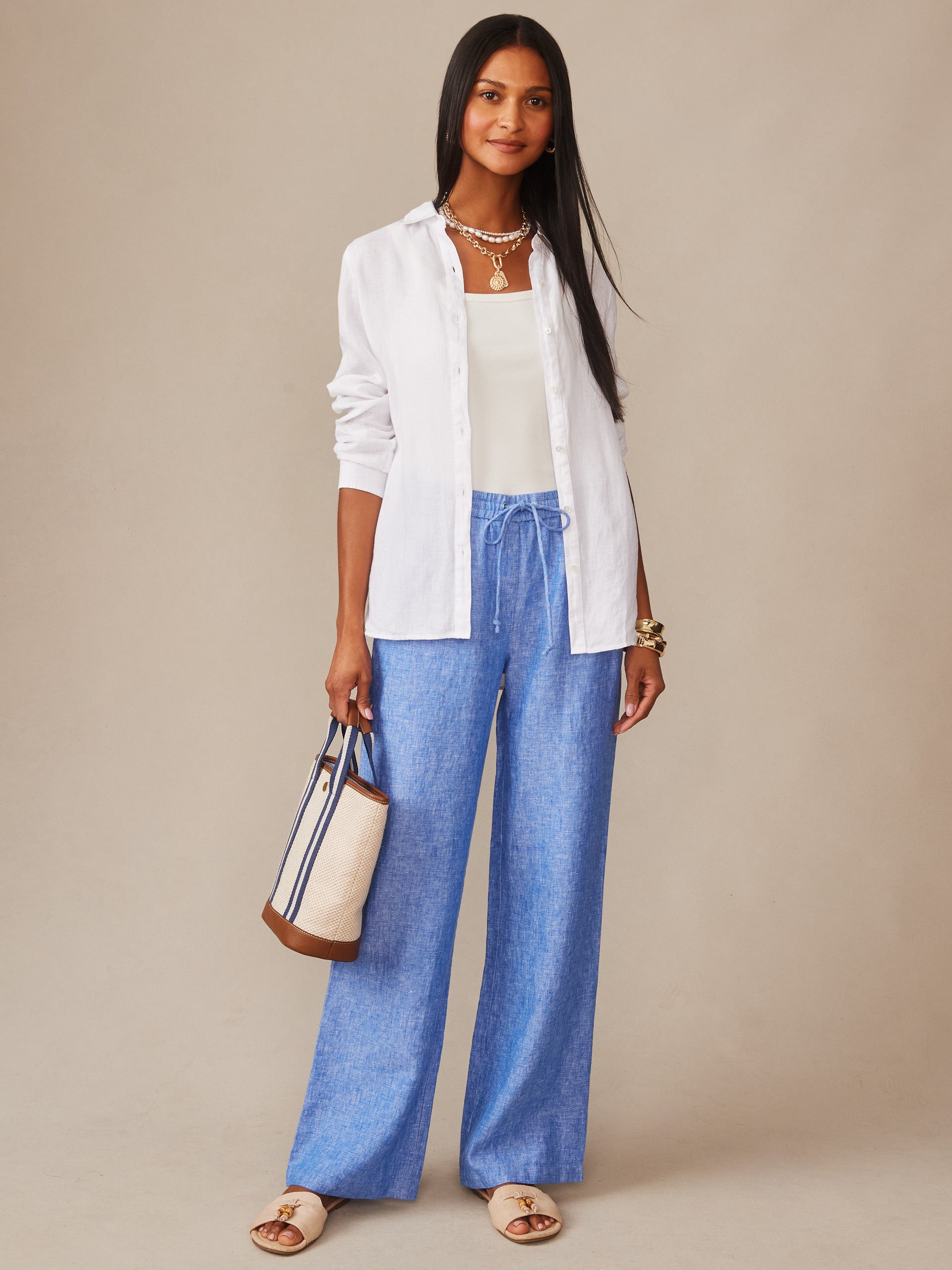 A stylish woman wearing J.McLaughlin's Irene Linen Pants in a relaxed fit paired with a white shirt and tank, carrying a chic tote, embodying effortlessly stylish, enduring designs.