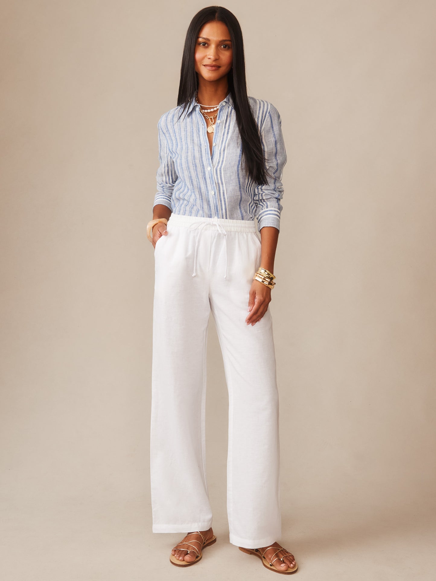 Woman wearing white Irene Linen Pants from J.McLaughlin paired with a striped blue button-up shirt and sandals, embodying effortlessly stylish, enduring designs with a legacy of timeless fashion.