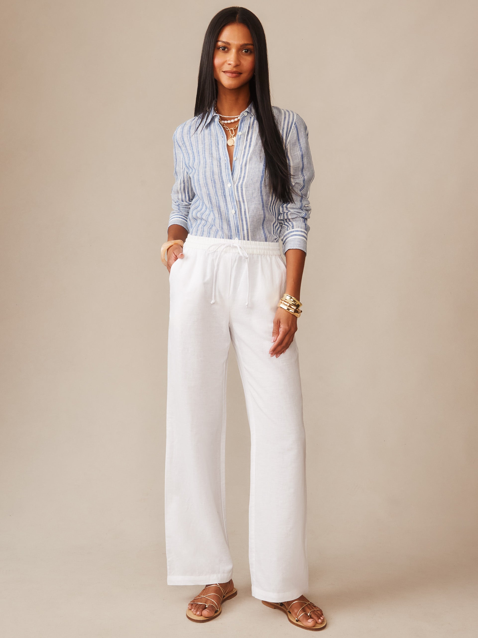 Woman wearing white Irene Linen Pants from J.McLaughlin paired with a striped blue button-up shirt and sandals, embodying effortlessly stylish, enduring designs with a legacy of timeless fashion.
