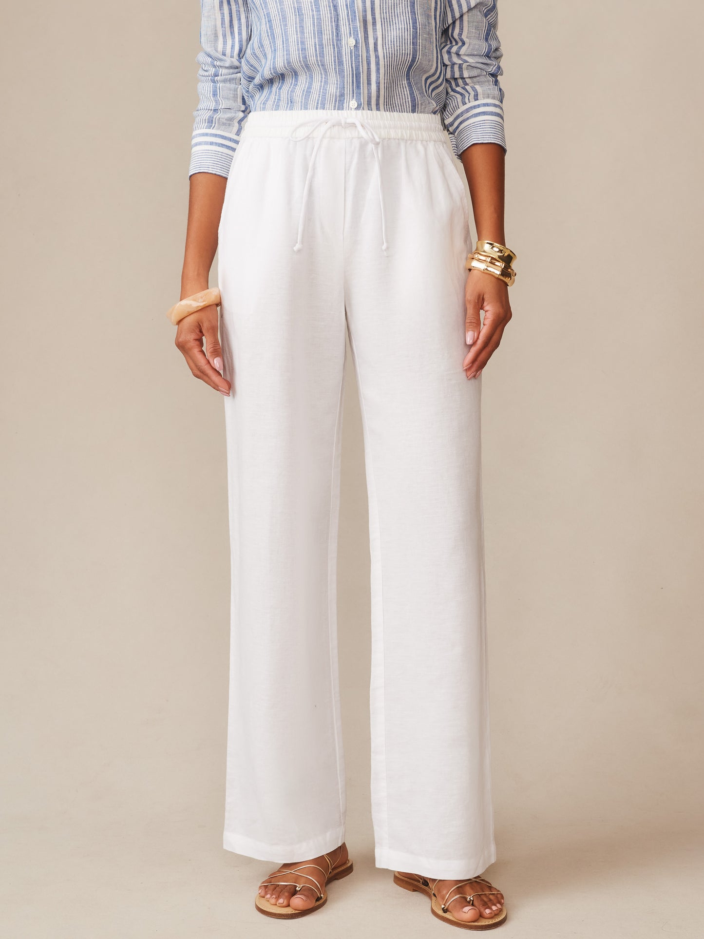 Front view of a woman wearing white Irene Linen Pants by J.McLaughlin paired with a striped blue shirt and gold bracelets, showcasing effortlessly stylish, enduring designs made with innovative fabrics.