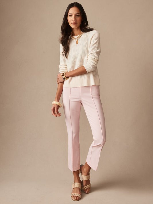 A woman models J.McLaughlin’s Ivy Pants in pale pink, paired with a textured cream sweater and layered gold jewelry, showcasing effortlessly stylish, enduring designs and innovative fabrics.