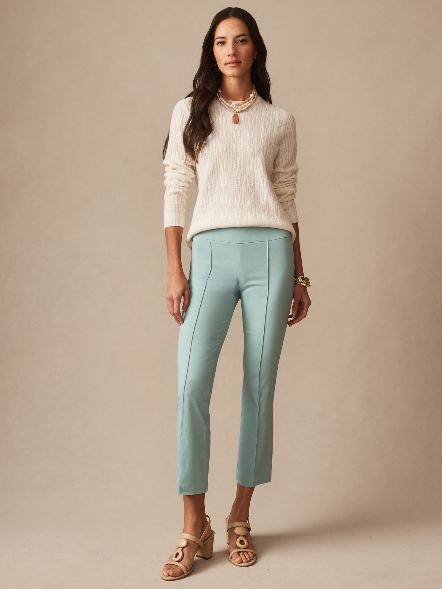 A woman wearing seafoam Ivy Pants by J.McLaughlin paired with a cream cable-knit sweater and stylish sandals, showcasing effortlessly stylish, enduring designs in innovative fabrics.