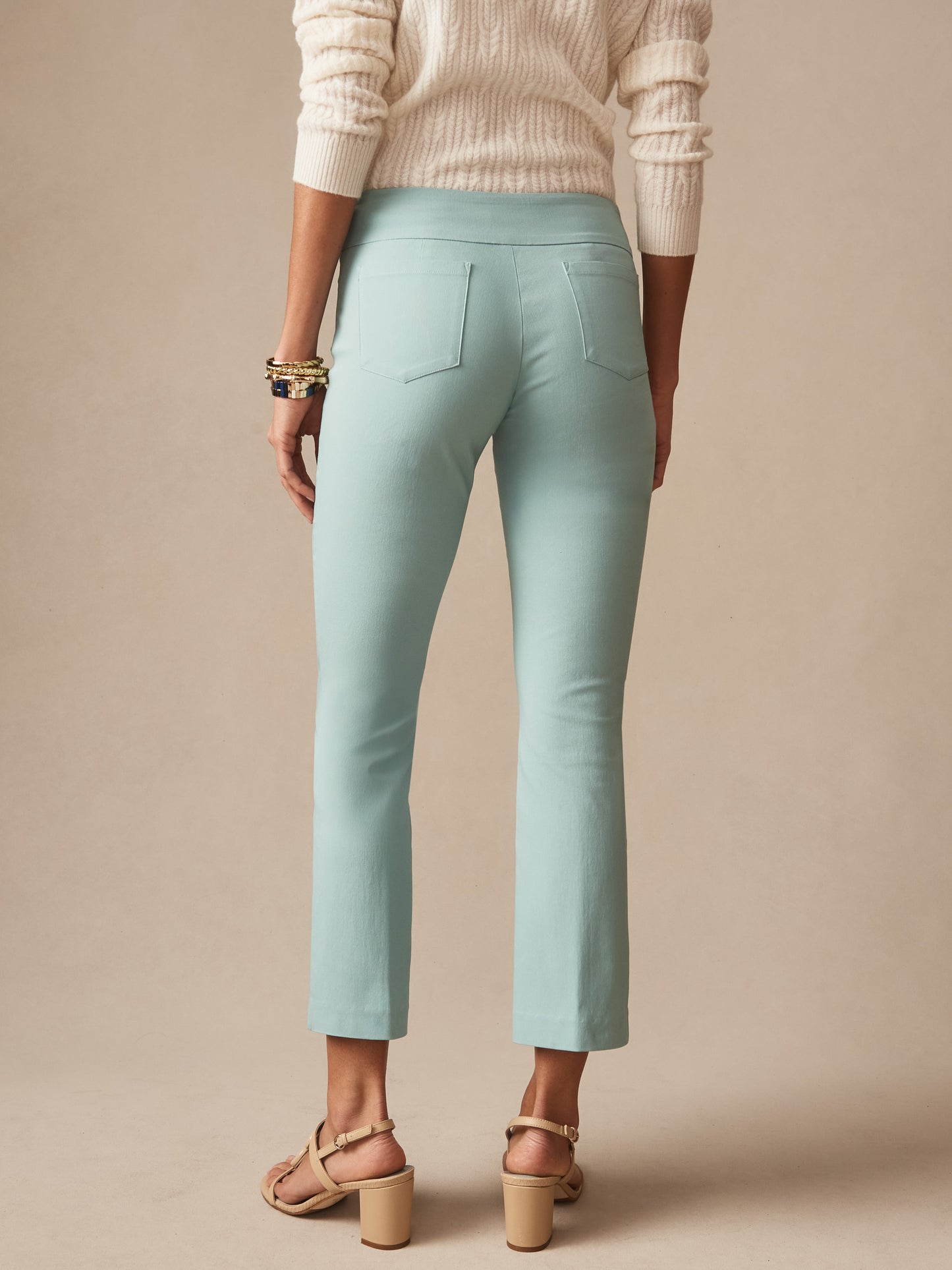 Back view of a woman wearing seafoam Ivy Pants by J.McLaughlin paired with a cream sweater and beige heeled sandals, showcasing effortlessly stylish, enduring designs in innovative fabrics.