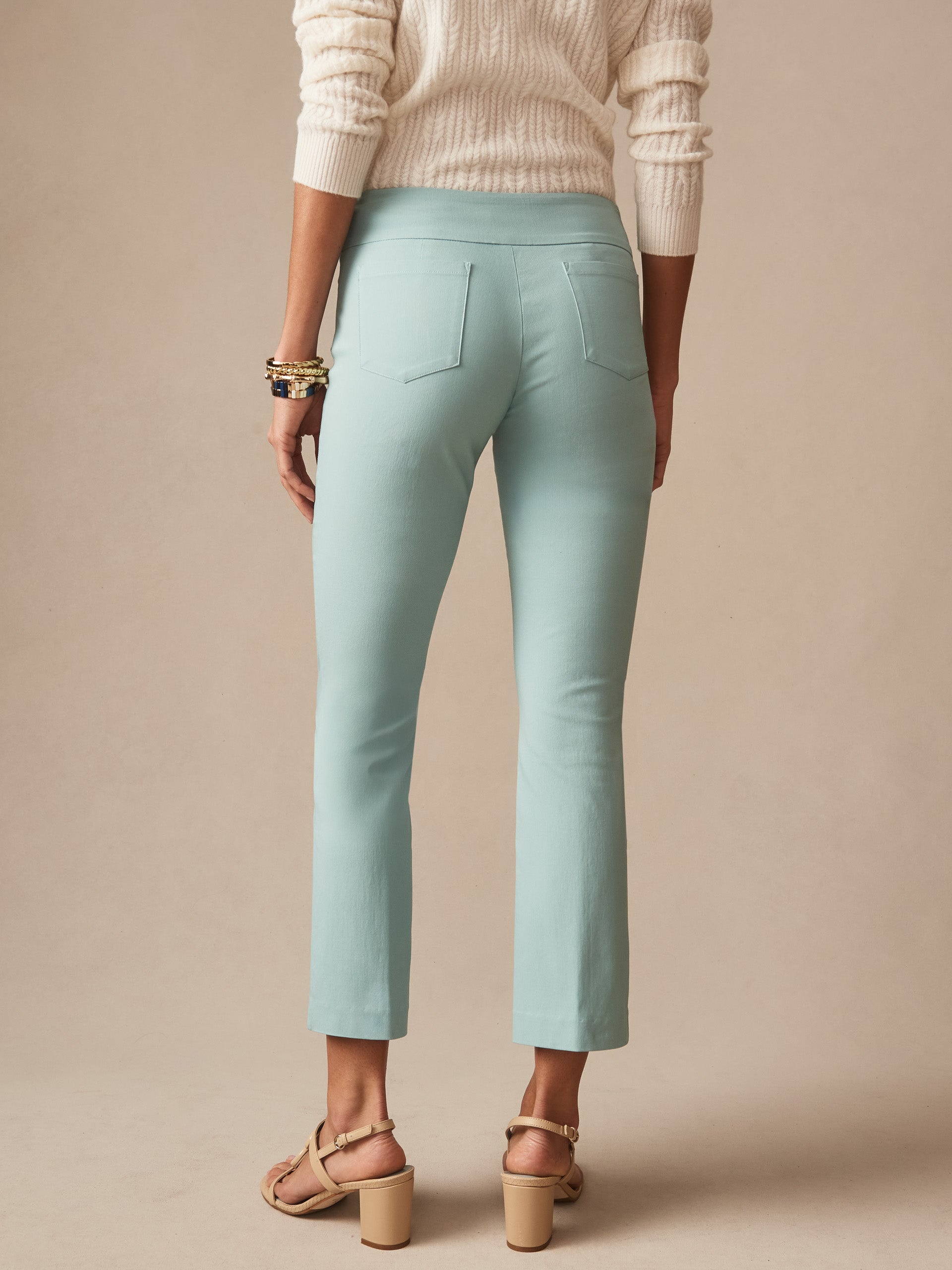 Back view of a woman wearing seafoam Ivy Pants by J.McLaughlin paired with a cream sweater and beige heeled sandals, showcasing effortlessly stylish, enduring designs in innovative fabrics.