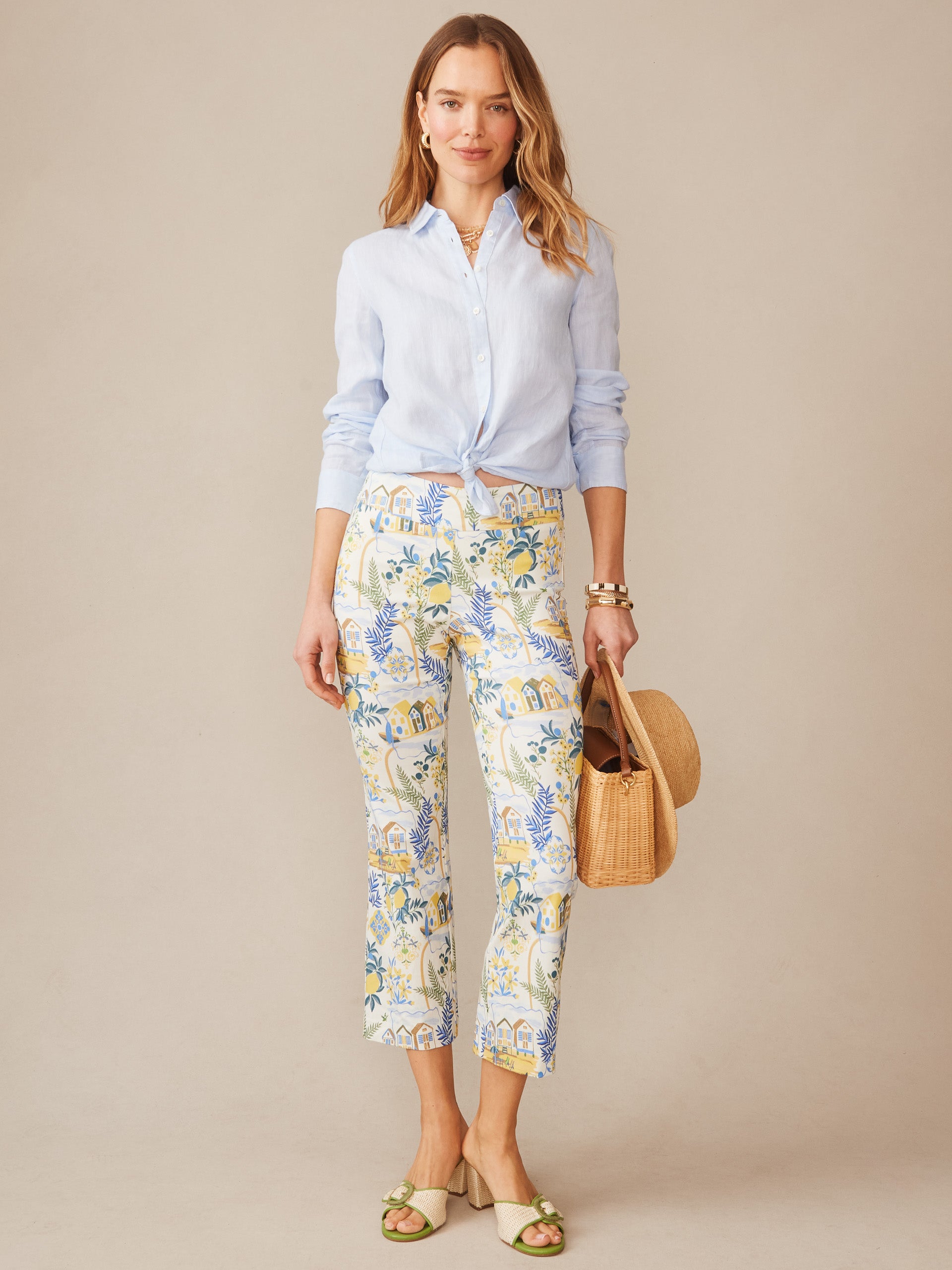 Woman wearing J.McLaughlin Ivy Pants in Amalfi Coast featuring a colorful coastal print paired with a light blue tied shirt, styled with sandals and a woven handbag, showcasing effortlessly stylish, enduring designs with a legacy of timeless fashion. A destination for defining style. Life, Well-Lived.