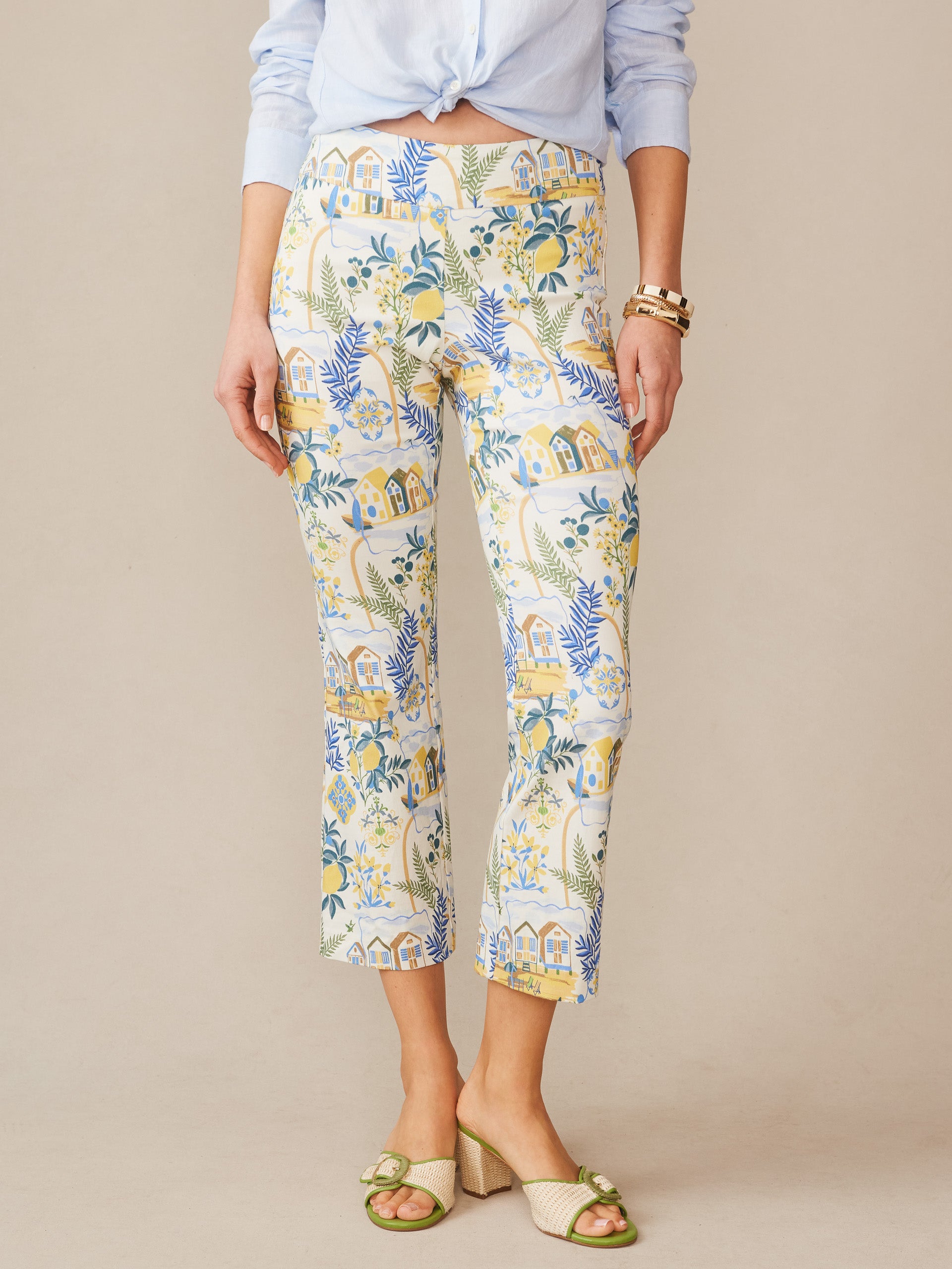 Woman wearing J.McLaughlin Ivy Pants in Amalfi Coast featuring an off-white background with blue, yellow, and green house and botanical print paired with a light blue shirt and stylish sandals, showcasing effortlessly stylish, enduring designs and innovative fabrics.
