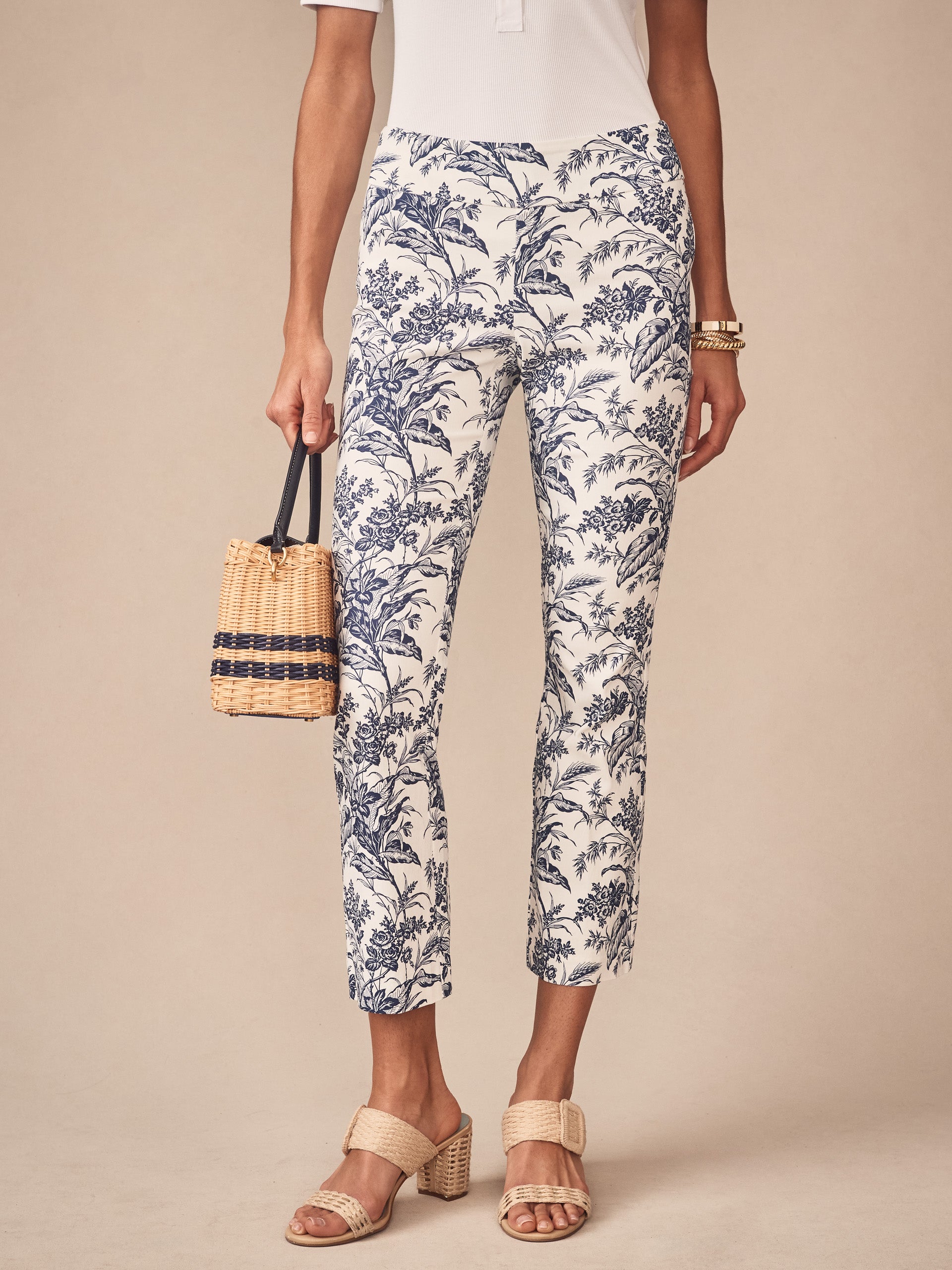 A model wearing J.McLaughlin Ivy Pants in Giardino Toile with a white and blue botanical print, paired with neutral heeled sandals and a woven handbag, showcasing effortlessly stylish, enduring designs crafted from innovative fabrics— a destination for defining style. Life, Well-Lived.