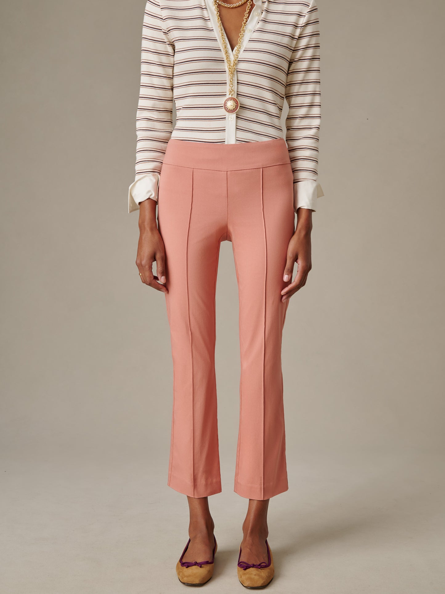 A model wearing J.McLaughlin Ivy Pants in a soft rose color paired with a striped blouse and layered gold necklaces, showcasing timeless style and impeccable craftsmanship.