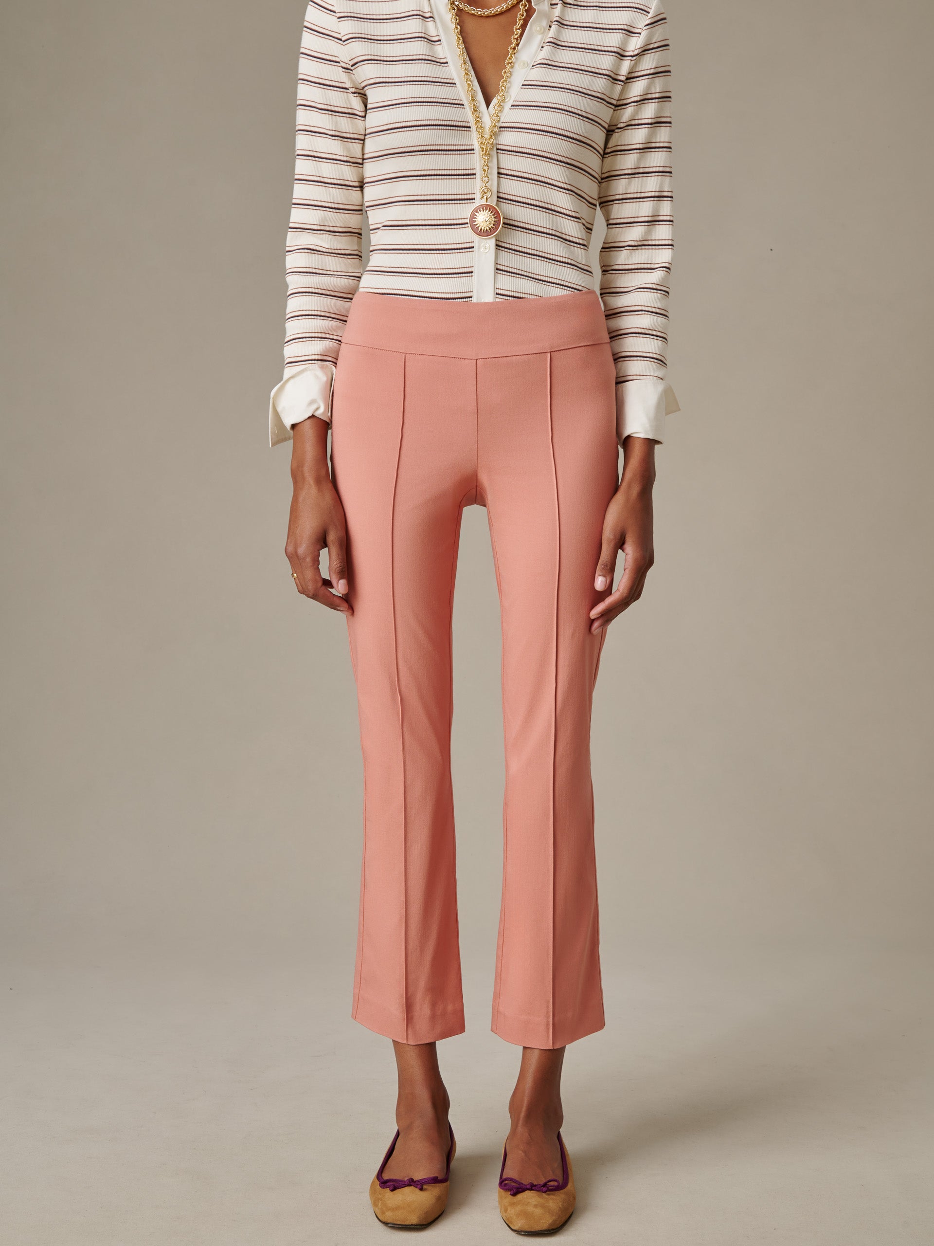 A model wearing J.McLaughlin Ivy Pants in a soft rose color paired with a striped blouse and layered gold necklaces, showcasing timeless style and impeccable craftsmanship.