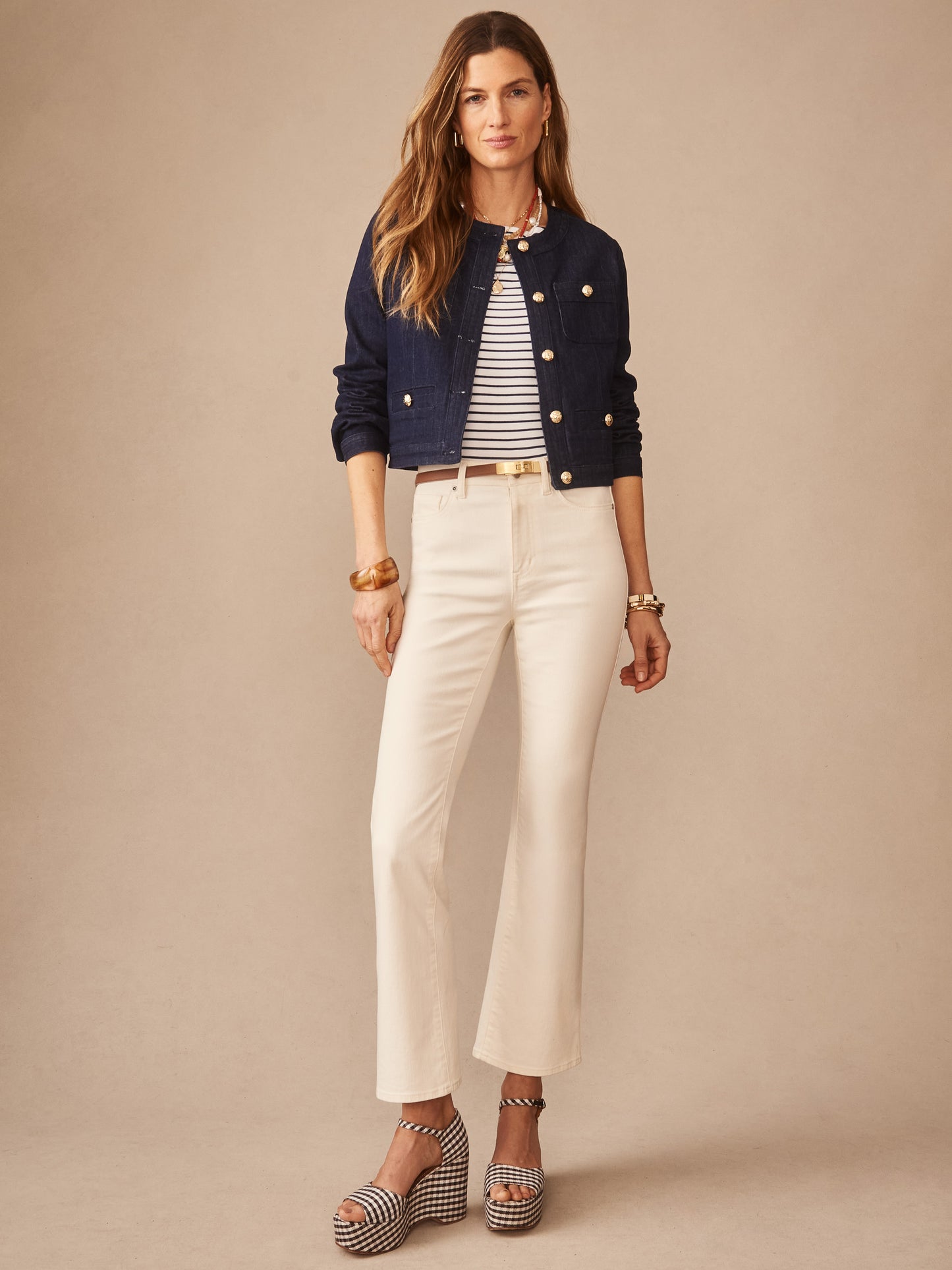 A woman models the Jackie Denim Jacket by J.McLaughlin in indigo, paired with cream pants, striped top, and platform sandals, showcasing effortlessly stylish, enduring designs.