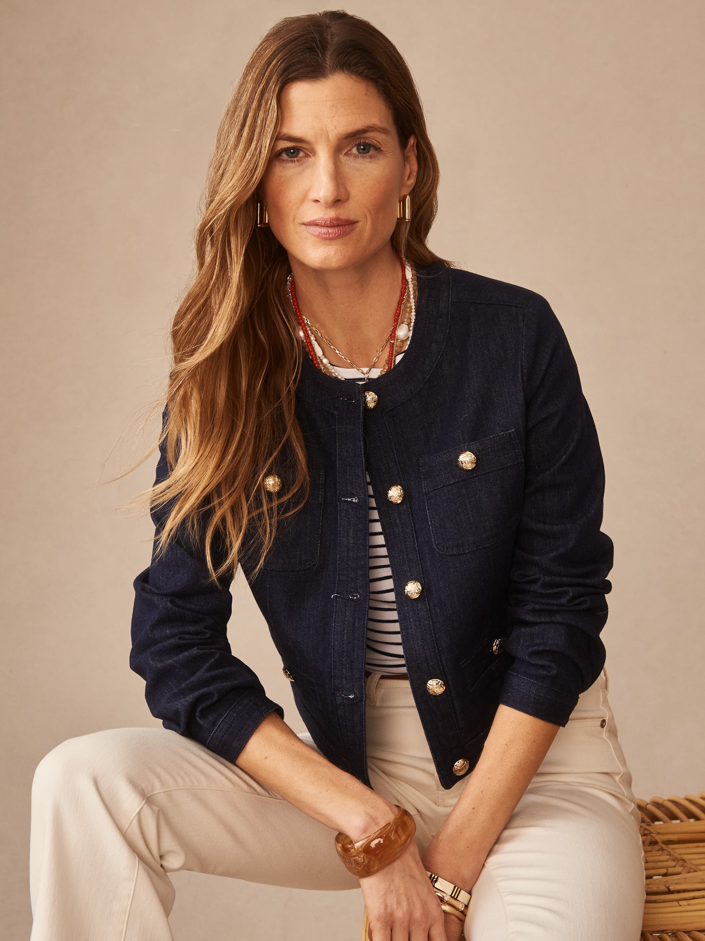 Woman wearing a Jackie Denim Jacket by J.McLaughlin in deep indigo, featuring gold buttons over a striped top and cream pants, embodying effortlessly stylish, enduring designs.