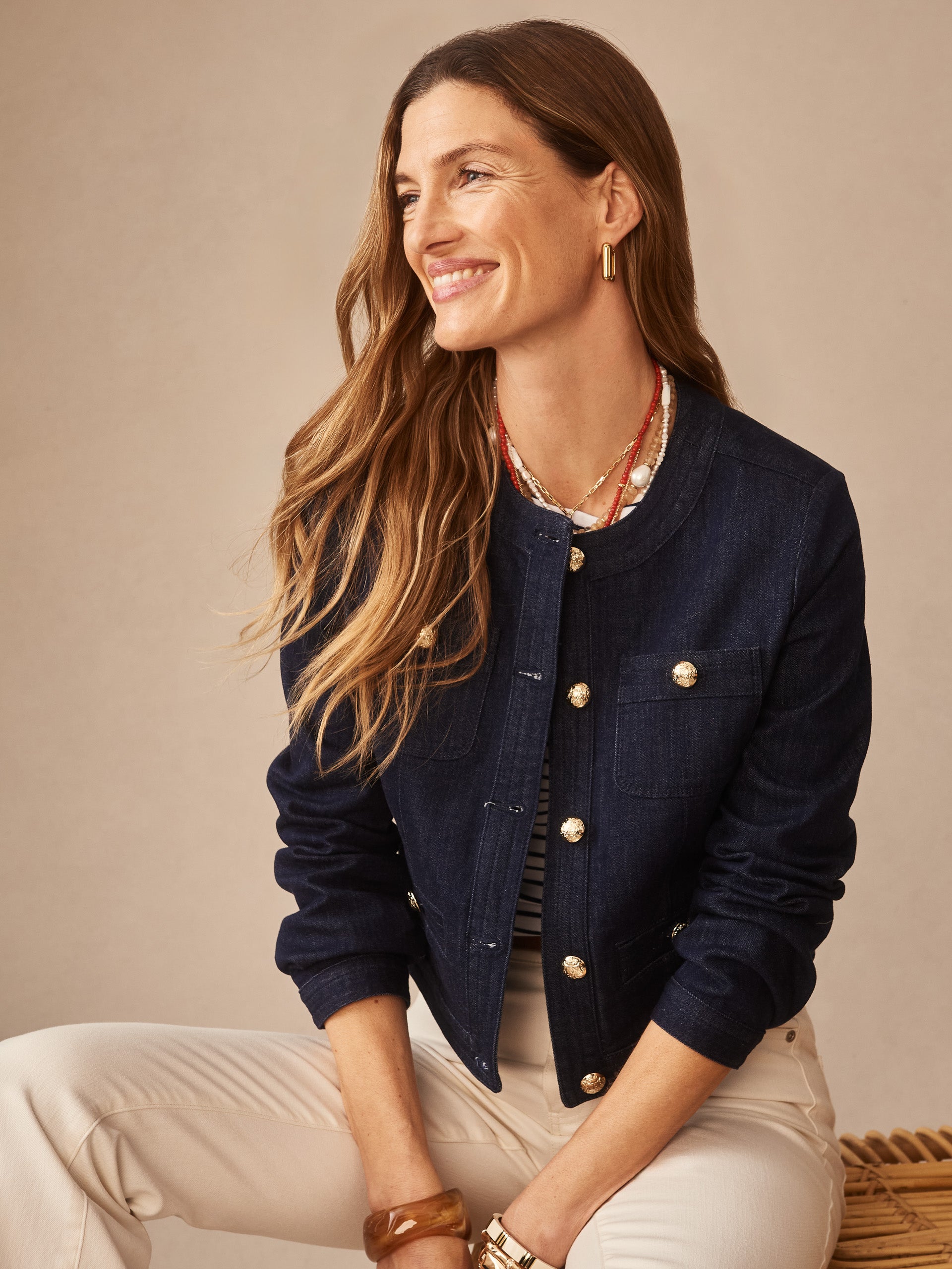 Woman smiling wearing a classic Jackie Denim Jacket by J.McLaughlin in indigo, paired with light-colored pants and layered necklaces, embodying effortlessly stylish, enduring designs and innovative fabrics.