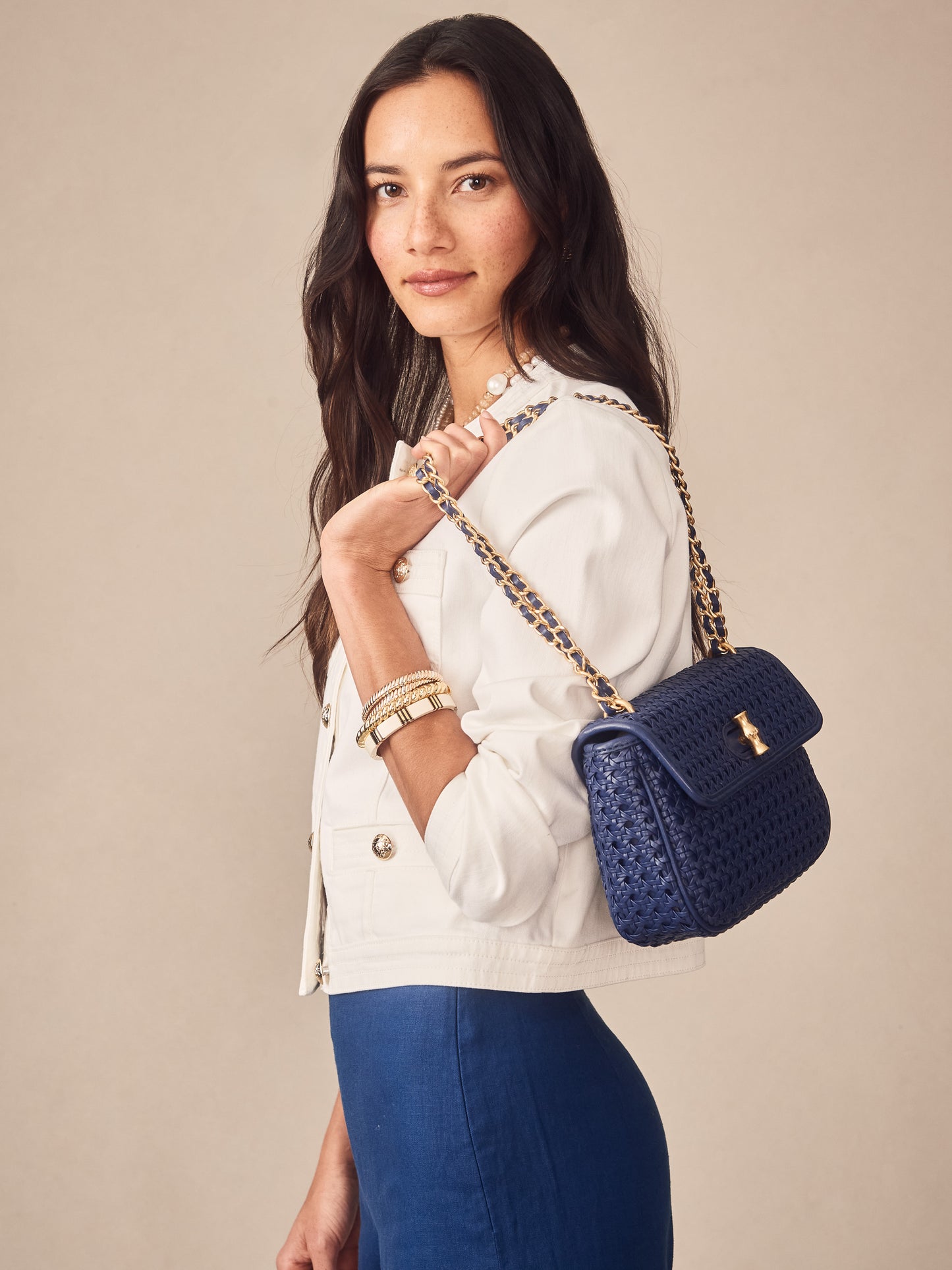 Woman wearing a white jacket and blue skirt carrying a navy Waverly Leather Handbag by J.McLaughlin with gold chain strap, showcasing effortlessly stylish, enduring designs.