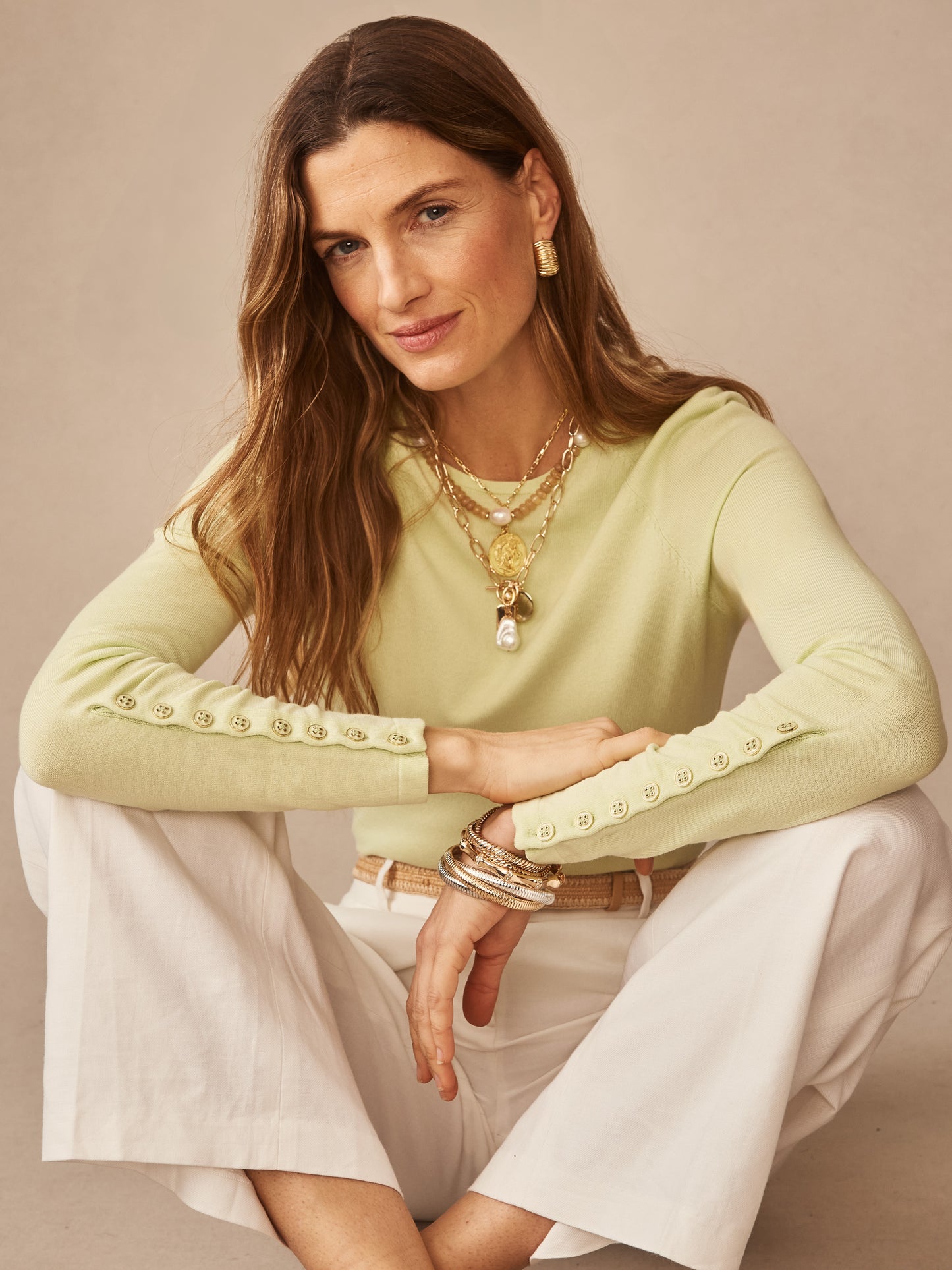 Woman wearing a lime green Jamey Sweater by J.McLaughlin, styled with layered gold necklaces and bracelets, sitting comfortably in white pants, embodying effortlessly stylish, enduring designs and innovative fabrics in a legacy of timeless fashion. A destination for defining style. Life, Well-Lived.