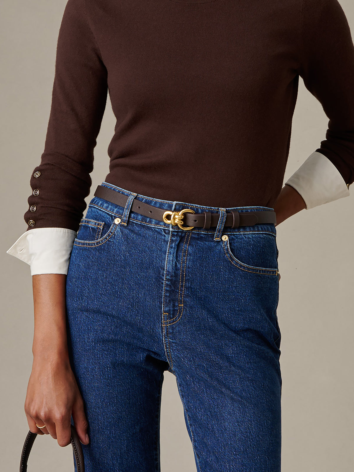 A close-up of a person wearing a brown Natalie Leather Belt by J.McLaughlin with gold knot hardware over blue denim jeans and a brown top with white cuffs, showcasing a timeless and effortlessly stylish look.