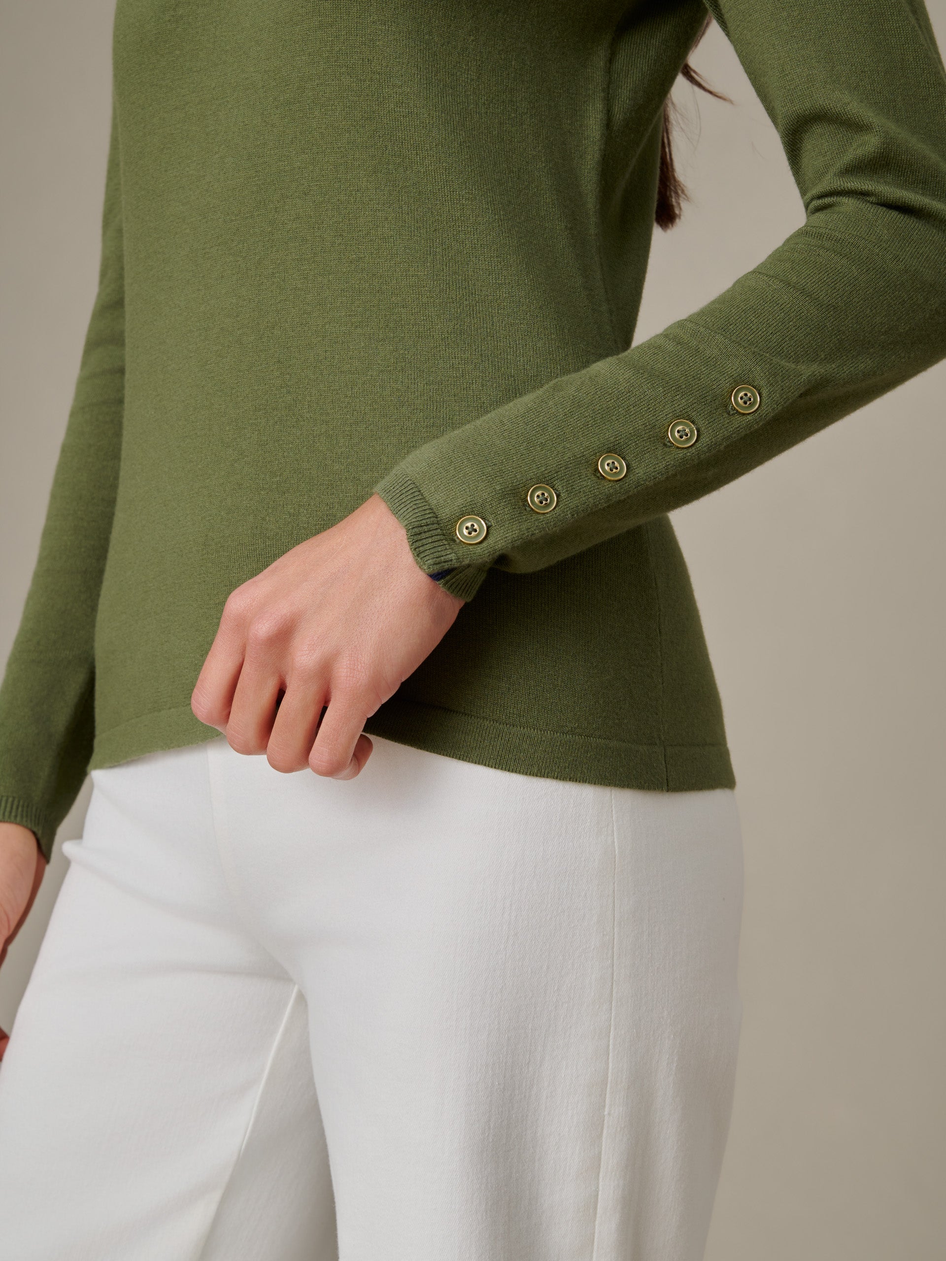 Close-up of a person wearing a Jamey Sweater in olive green by J.McLaughlin, featuring button details on the sleeve, styled with white pants for a timeless, effortlessly stylish look.