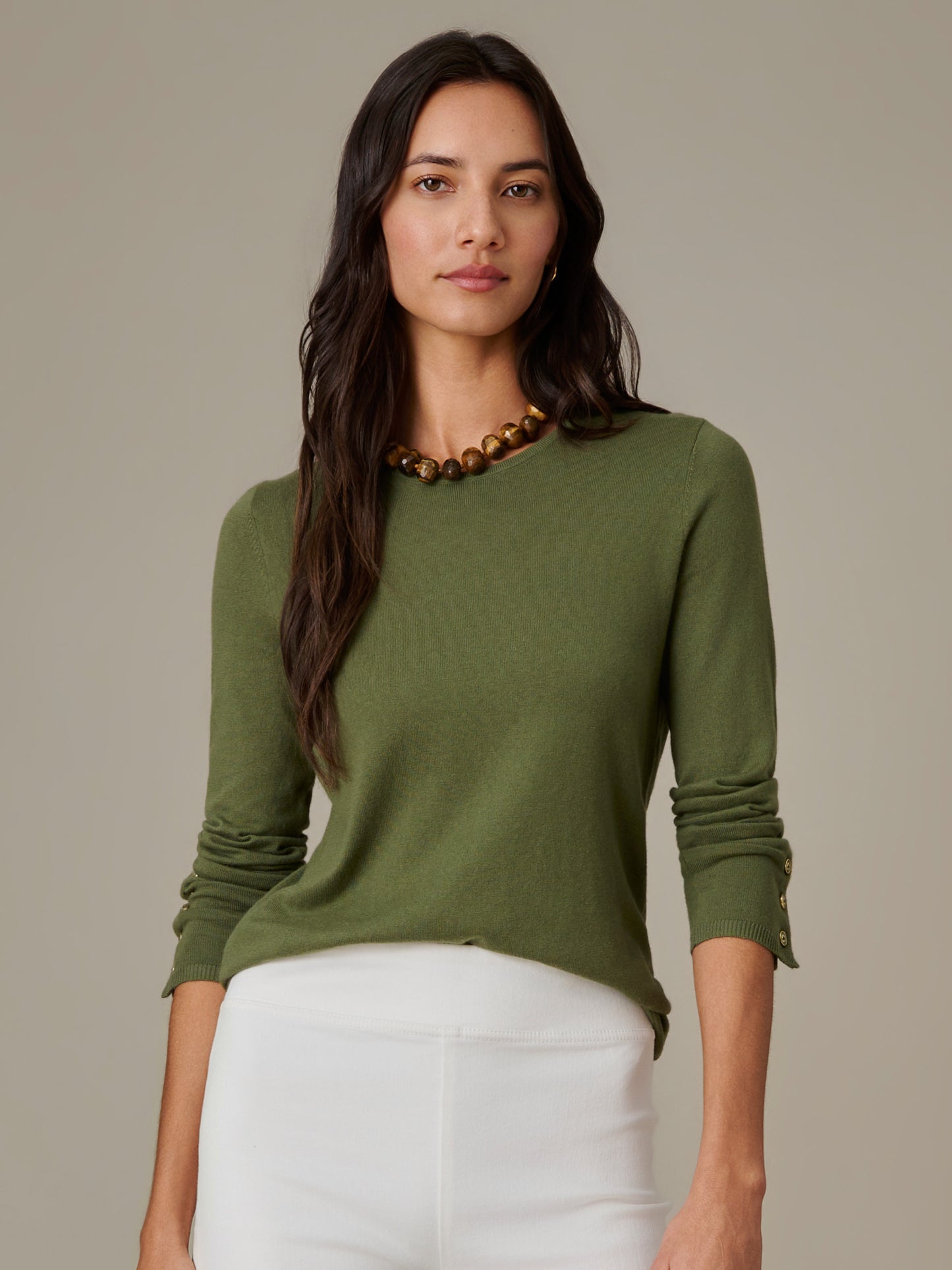 Woman wearing an olive green Jamey Sweater by J.McLaughlin, paired with white pants and a gold bead necklace, showcasing effortlessly stylish and enduring design.