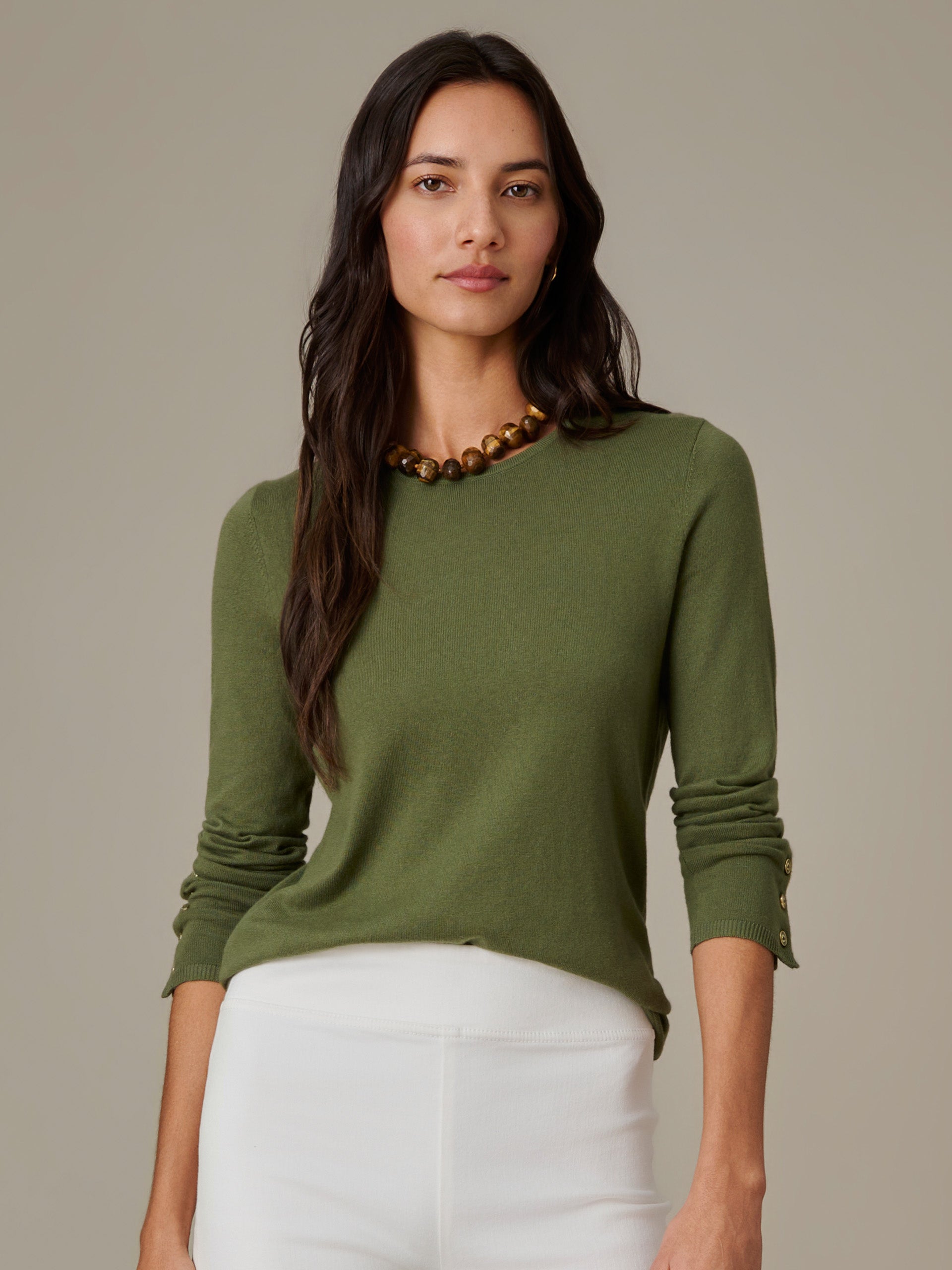 Woman wearing an olive green Jamey Sweater by J.McLaughlin, paired with white pants and a gold bead necklace, showcasing effortlessly stylish and enduring design.