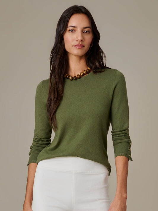Woman wearing an olive green Jamey Sweater by J.McLaughlin, paired with white pants and a gold bead necklace, showcasing effortlessly stylish and enduring design.