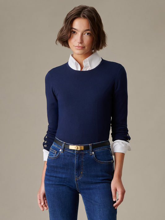 A woman wearing a navy Jamey Sweater over a white button-down shirt paired with dark blue jeans and a black belt, showcasing impeccable craftsmanship from J.McLaughlin.