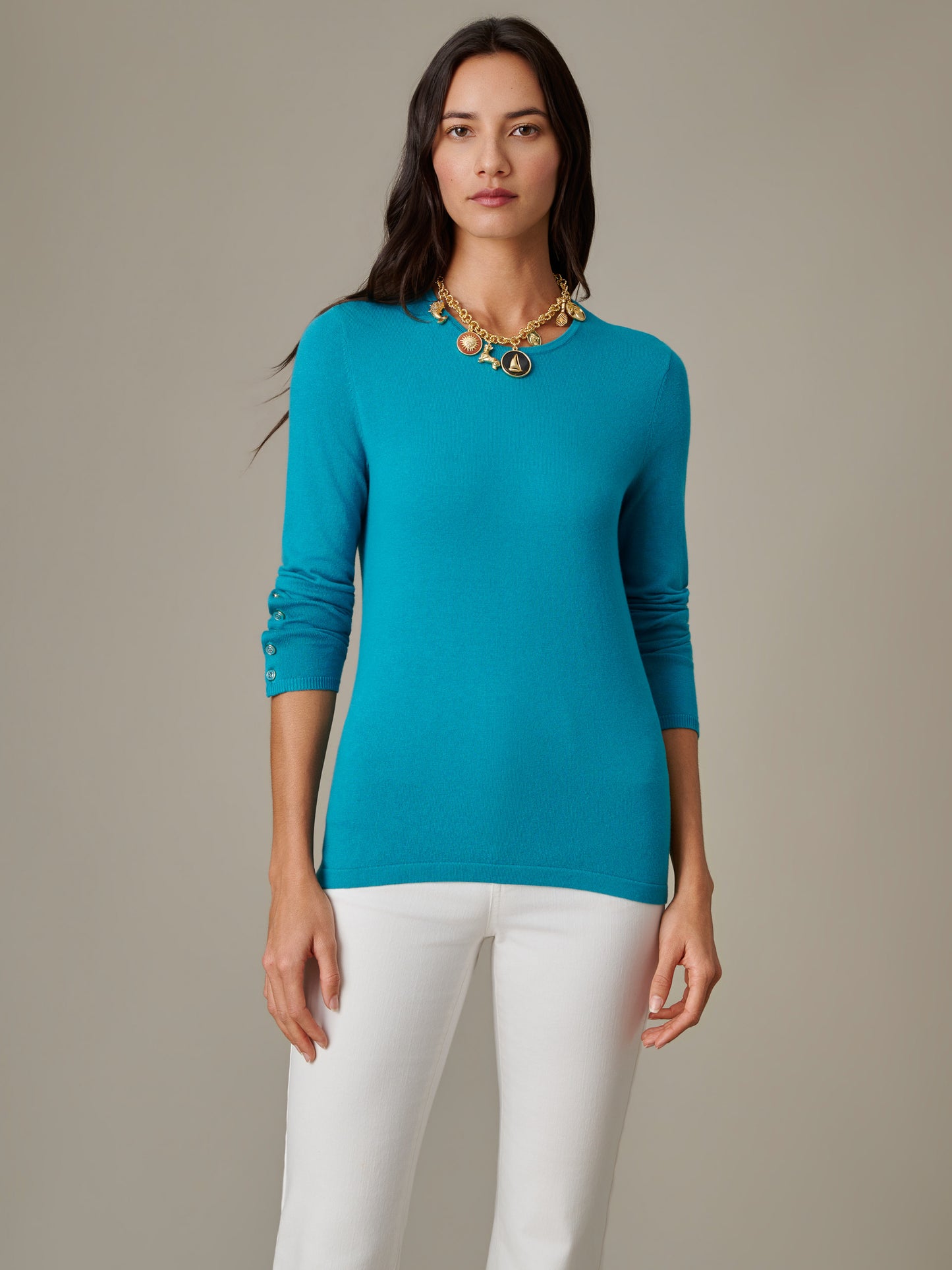 Woman wearing a vibrant blue Jamey Sweater by J.McLaughlin paired with white pants and a gold statement necklace, showcasing effortlessly stylish, timeless design.