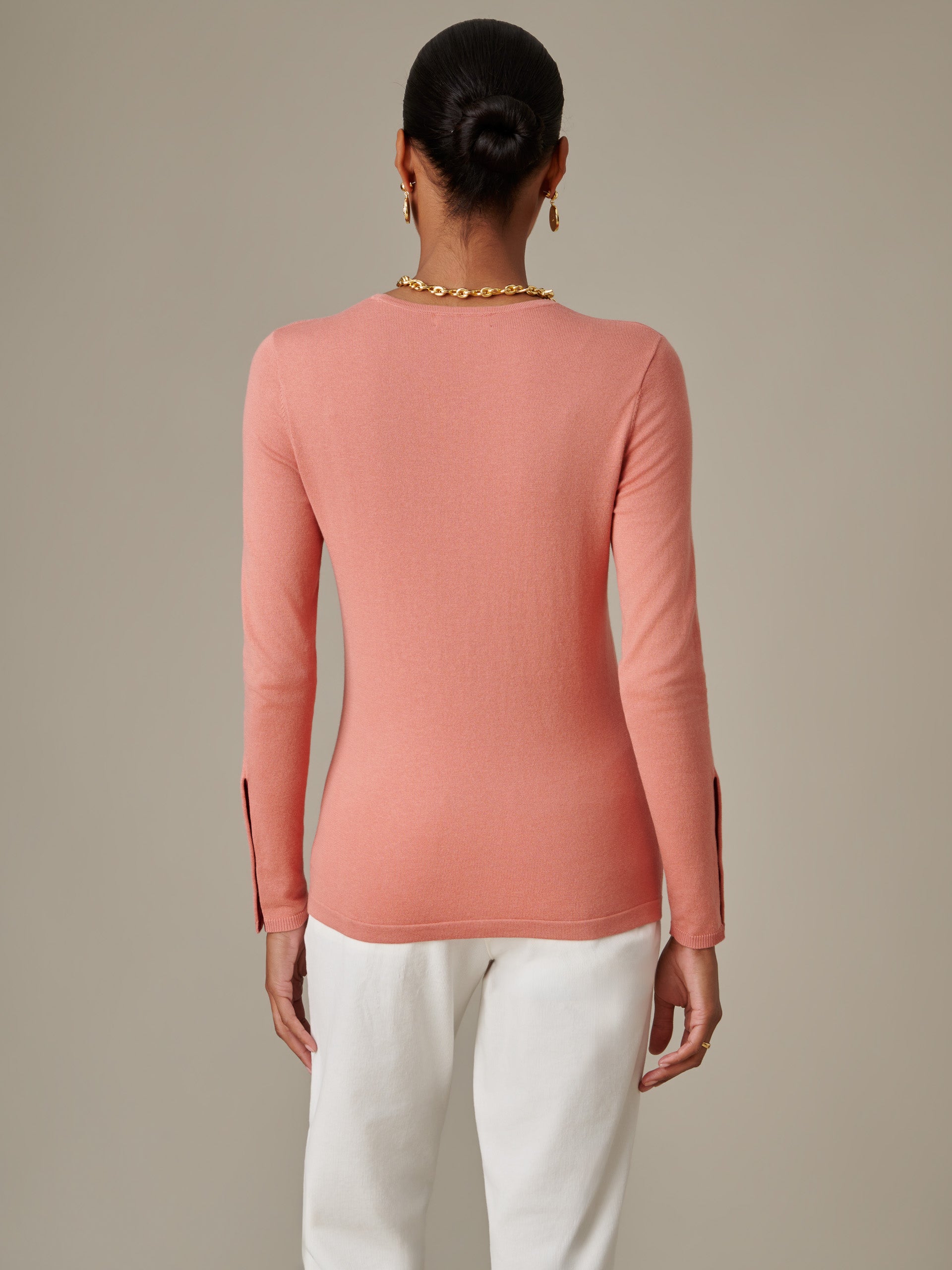 Woman wearing a fitted long-sleeve pink Jamey Sweater by J.McLaughlin with slits at the cuffs, paired with white pants, showcasing timeless style and craftsmanship.