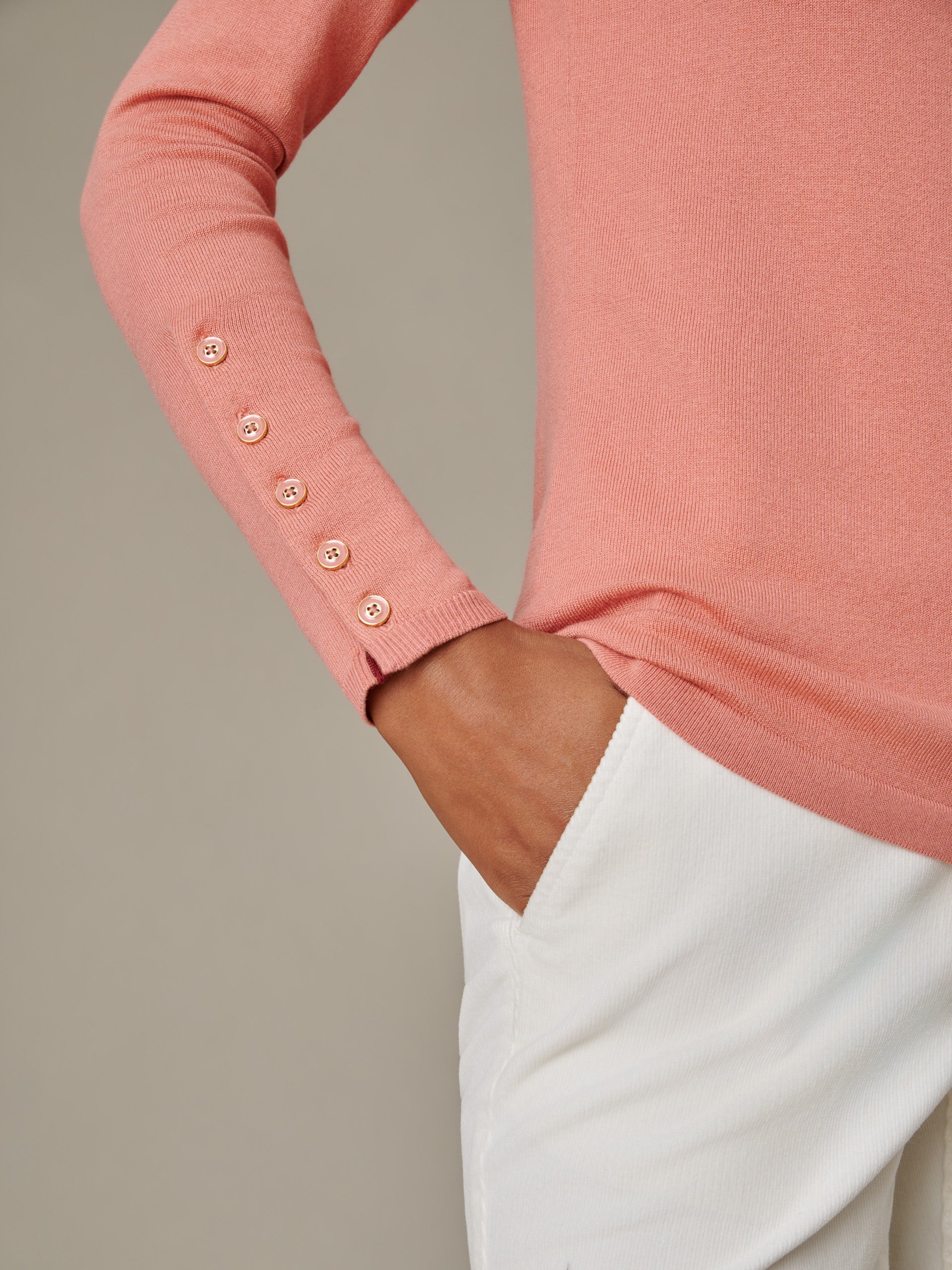 Close-up of a soft coral Jamey Sweater sleeve with four buttons near the cuff, styled with white pants, showcasing J.McLaughlin's effortlessly stylish and enduring design.