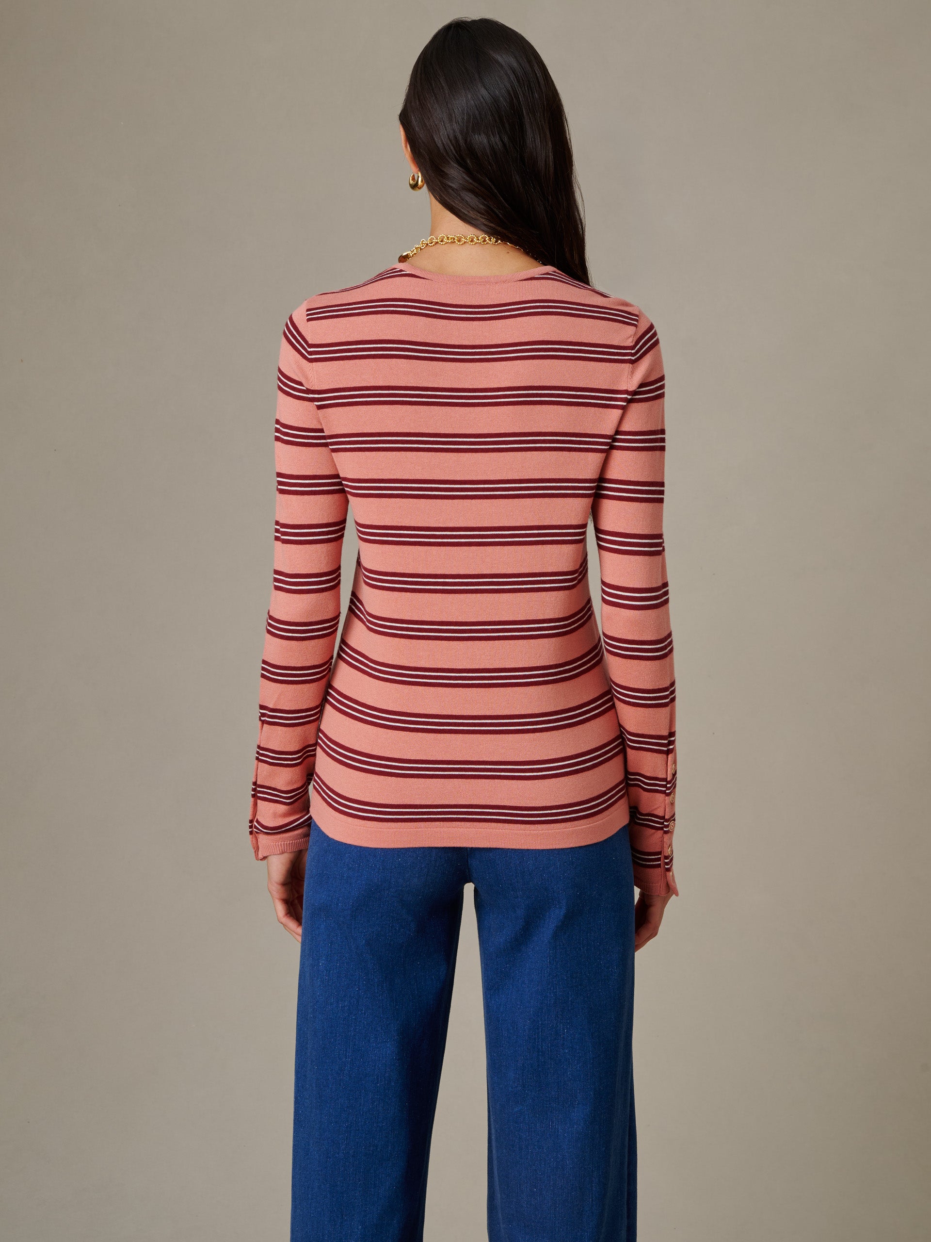 Woman seen from behind wearing a J.McLaughlin Jamey Sweater in Stripe with horizontal burgundy stripes on a muted pink background, paired with blue pants in a simple, timeless style.