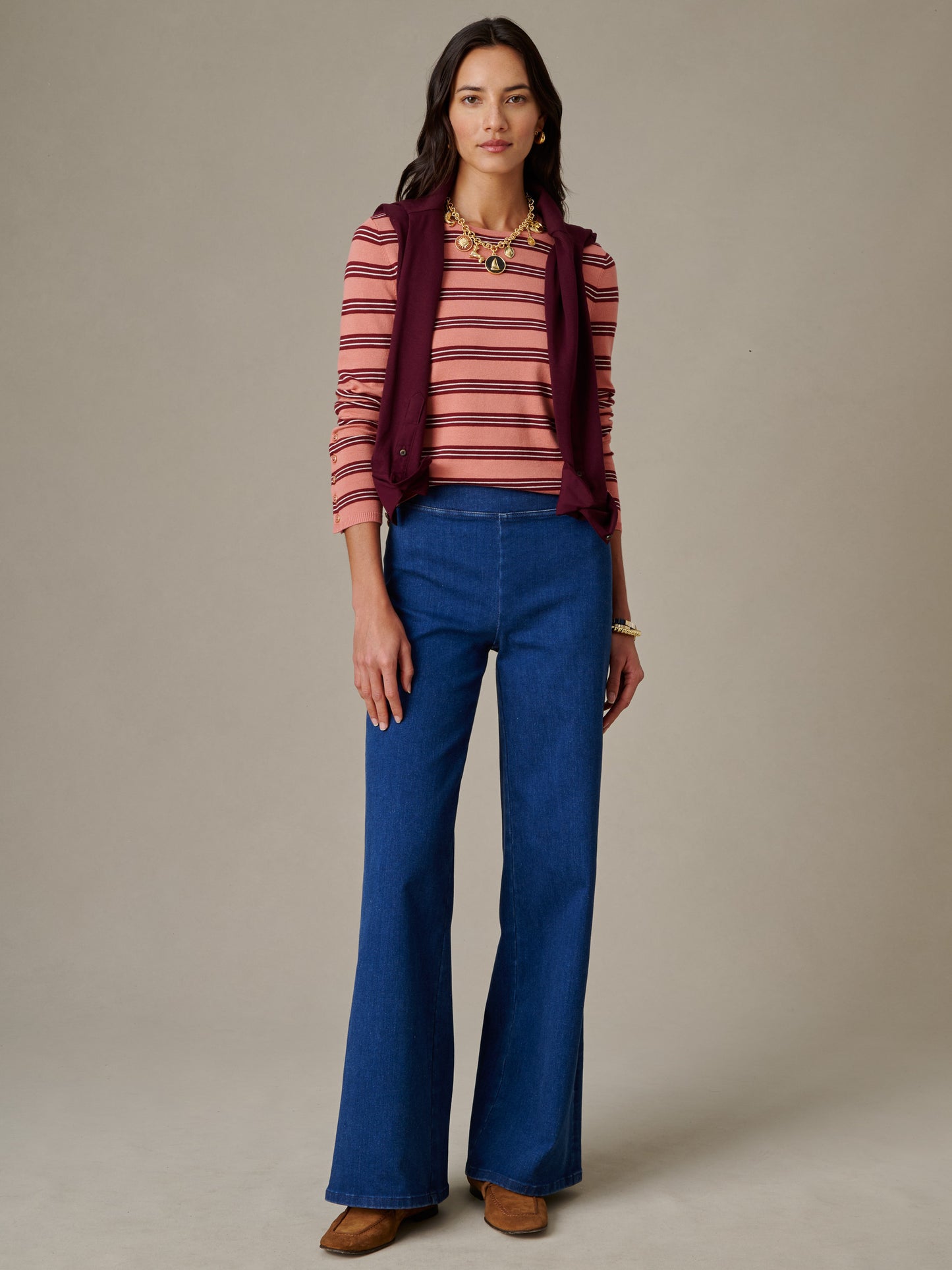Woman wearing J.McLaughlin Jamey Sweater in Stripe paired with blue wide-leg jeans and a sleeveless burgundy vest, styled with gold jewelry and brown shoes.