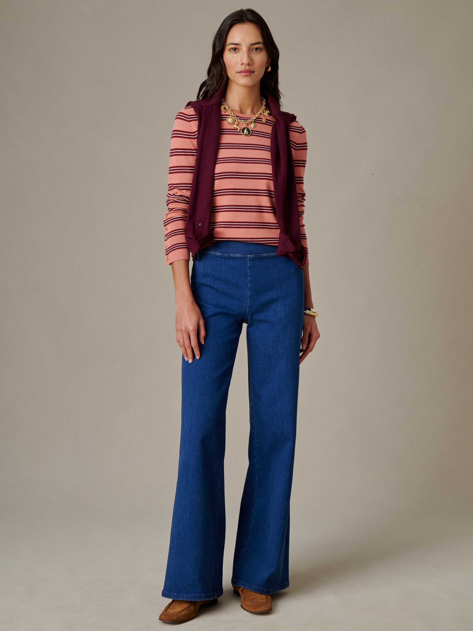 Woman wearing J.McLaughlin Jamey Sweater in Stripe paired with blue wide-leg jeans and a sleeveless burgundy vest, styled with gold jewelry and brown shoes.