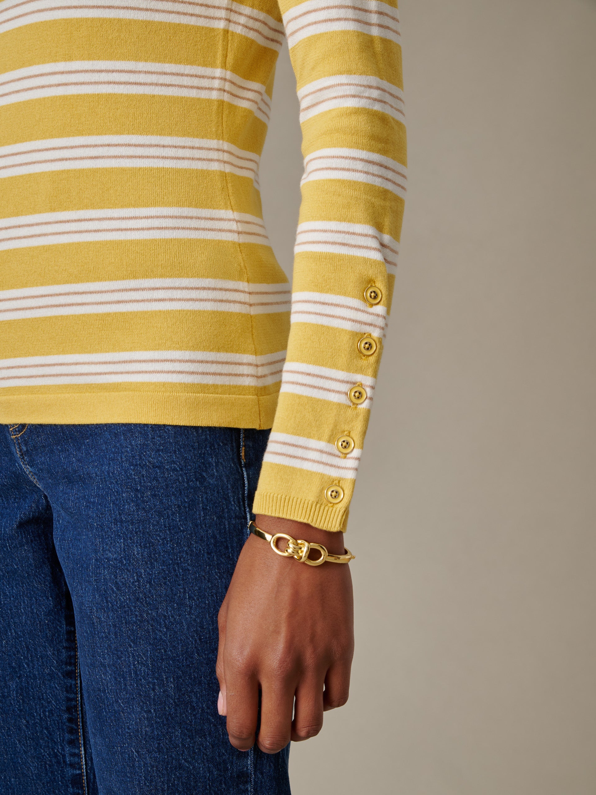 Close-up of an arm wearing the Jamey Sweater in Stripe by J.McLaughlin, featuring yellow and white stripes with decorative buttons on the cuff, paired with dark denim jeans and a gold bracelet, showcasing effortlessly stylish, enduring design.