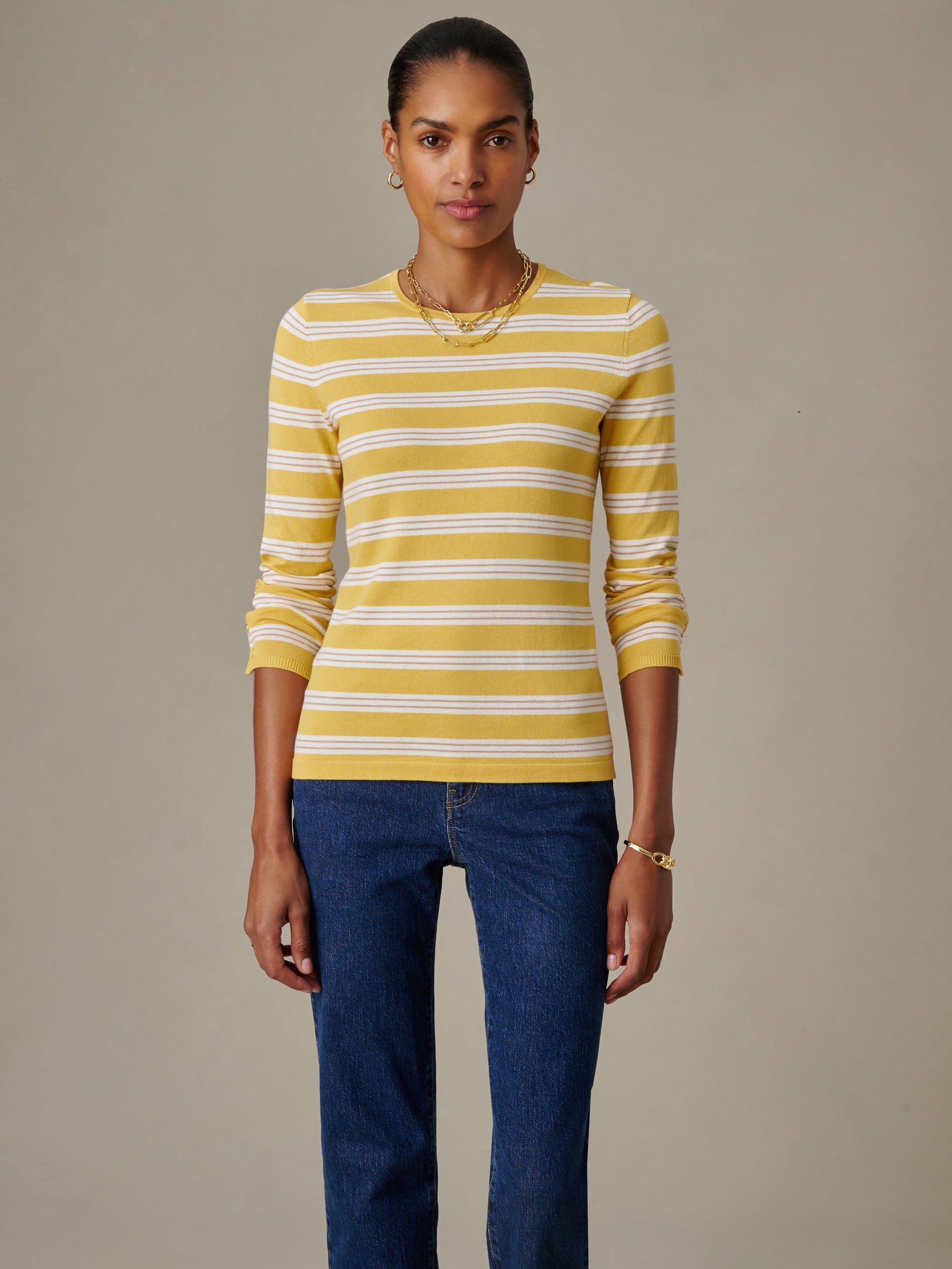 A woman wearing the Jamey Sweater in Stripe by J.McLaughlin in yellow and white stripes, paired with blue jeans, showcasing effortlessly stylish and enduring designs.