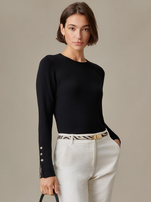 Woman wearing a black Jamey Sweater by J.McLaughlin featuring tailored set-in sleeves and signature enamel buttons, paired with white pants and a patterned belt, embodying timeless style.