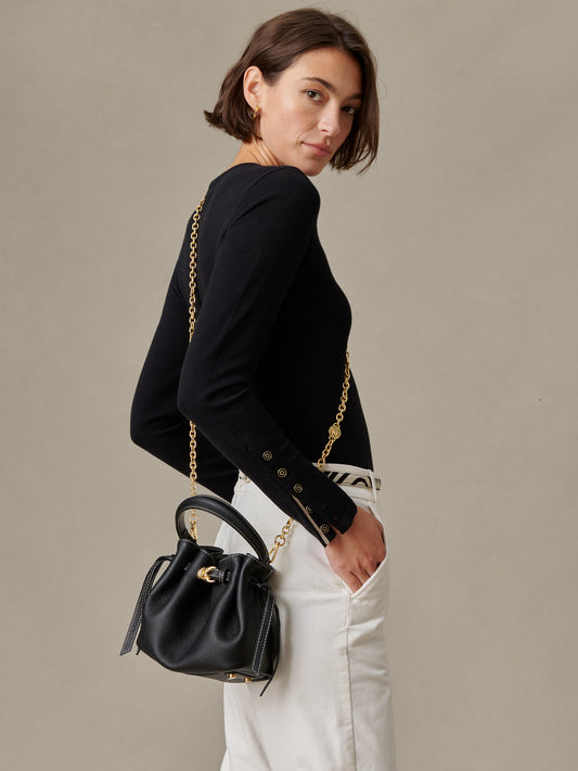 Woman wearing a black long-sleeve top and white pants, carrying The Mini Lex Leather Handbag by J.McLaughlin with a gold chain strap over her shoulder, showcasing an effortlessly stylish bag.