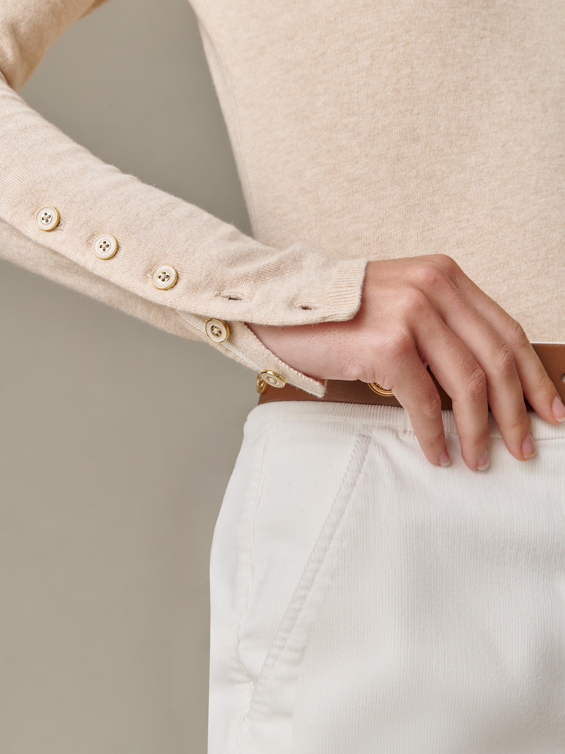Close-up of a hand adjusting a beige Jamey Sweater sleeve with button details, styled with white pants and a brown belt by J.McLaughlin, embodying timeless fashion and craftsmanship.