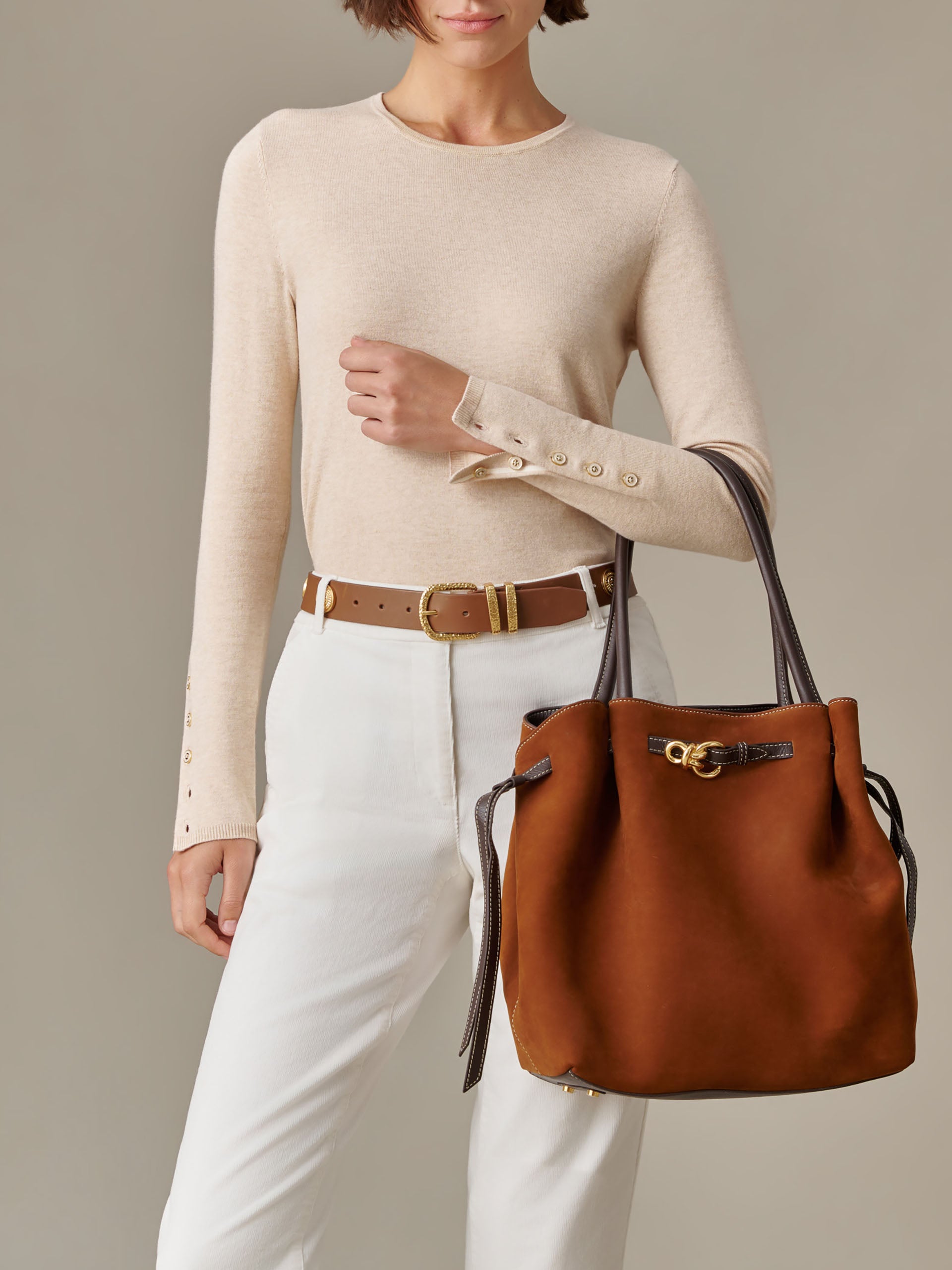 A model wearing a beige long sleeve top and white pants with a brown belt, holding The Lex Suede Tote Bag by J.McLaughlin in rich brown suede with dark brown handles and gold hardware, showcasing a blend of timeless style and craftsmanship.