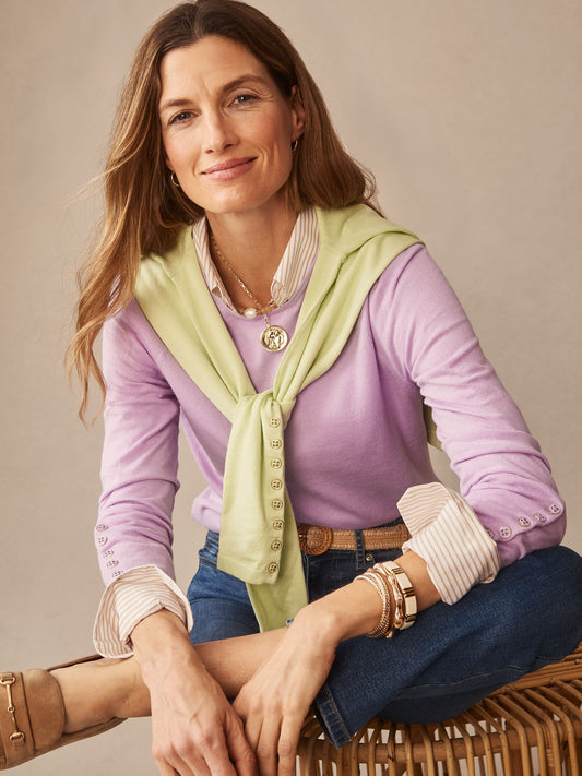A woman wearing a lavender Jamey Sweater layered over a striped shirt, with a light green sweater draped over her shoulders, styled with denim jeans and gold accessories, reflecting J.McLaughlin’s effortlessly stylish and enduring designs.