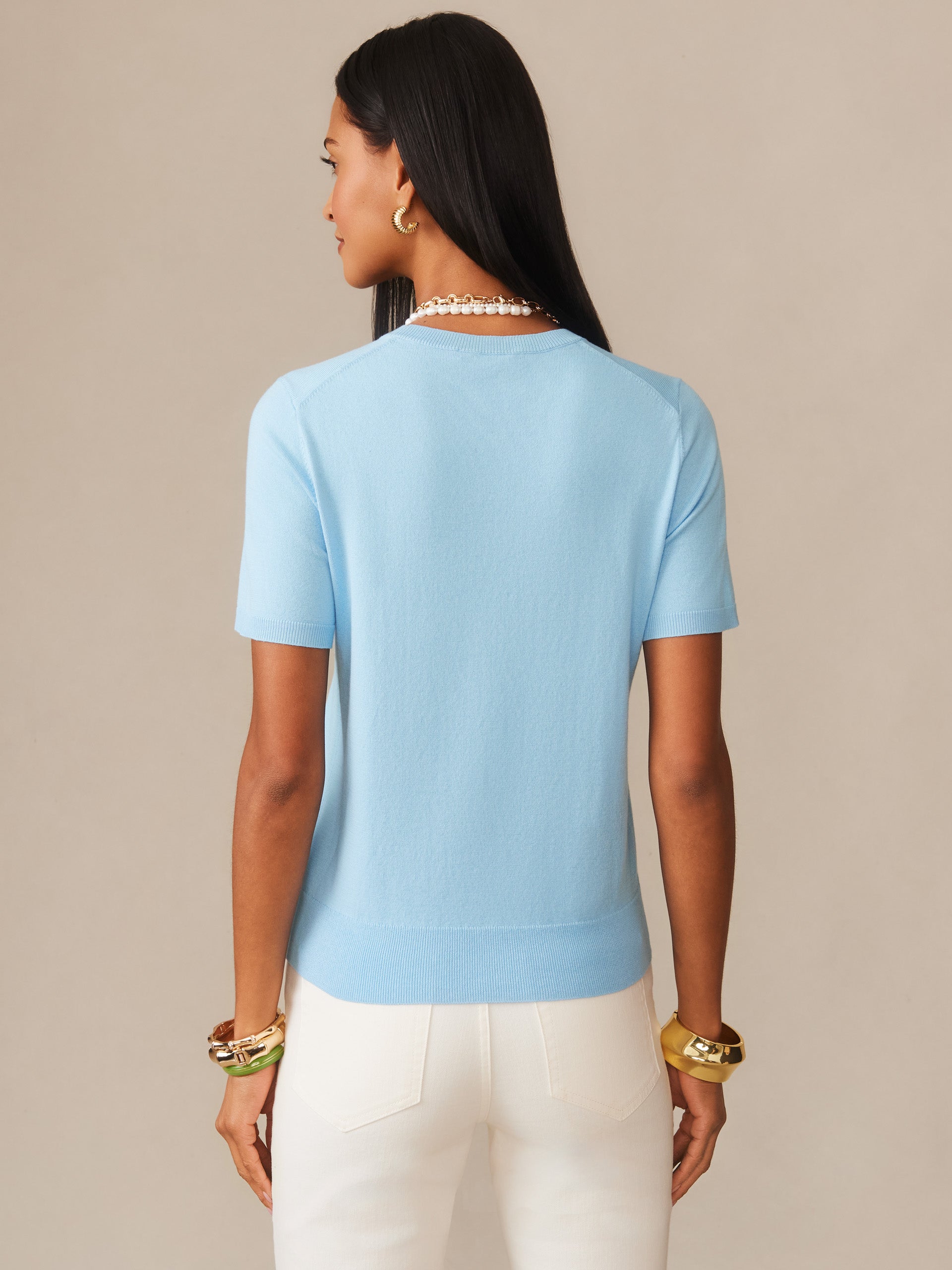 Woman wearing a baby blue Joelle Sweater by J.McLaughlin, styled with white pants and gold accessories, showcasing effortlessly stylish, enduring designs and innovative fabrics.