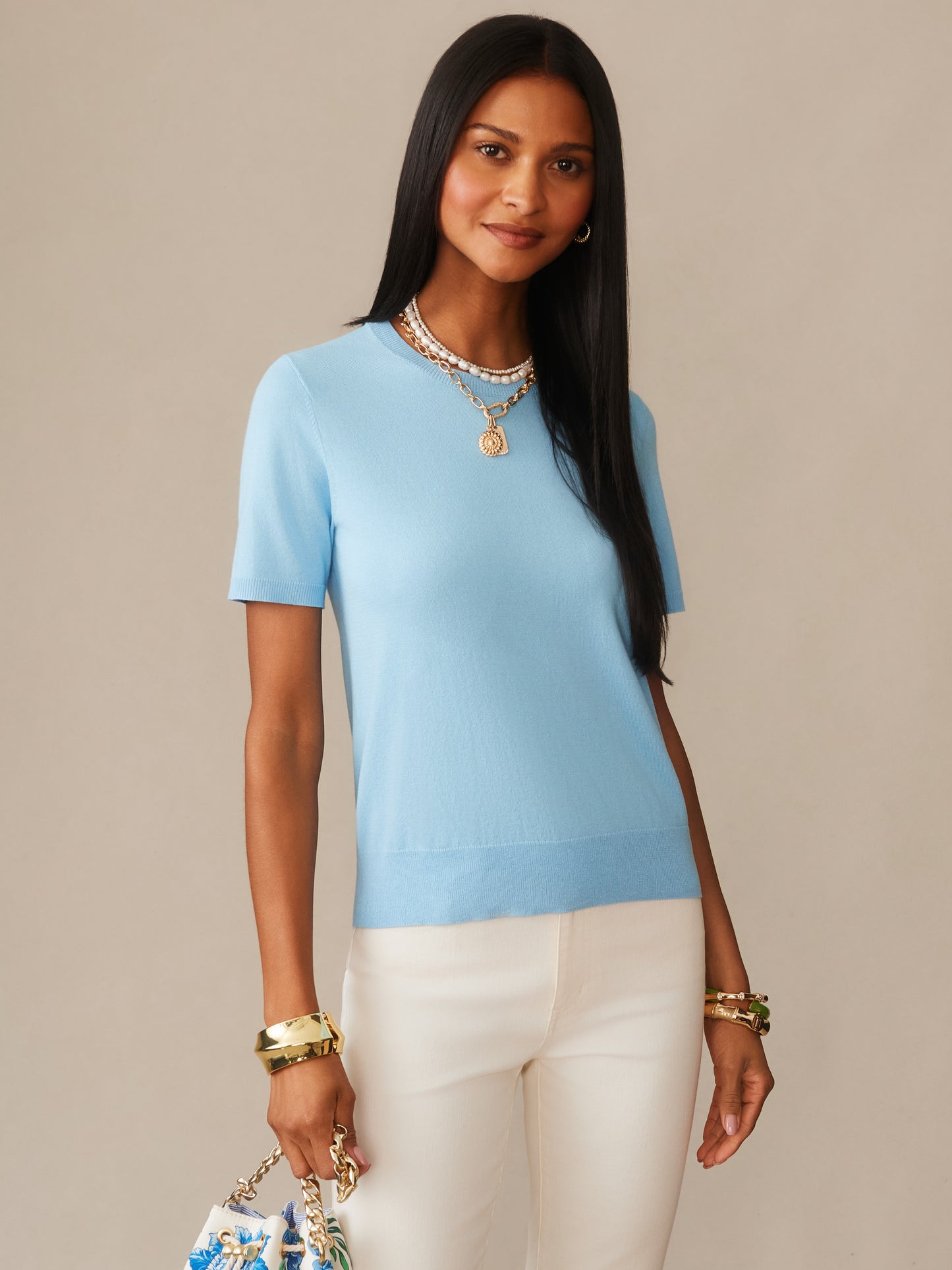 A woman wearing a baby blue Joelle Sweater by J.McLaughlin paired with white pants and layered gold necklaces, showcasing effortlessly stylish, enduring designs and innovative fabrics.