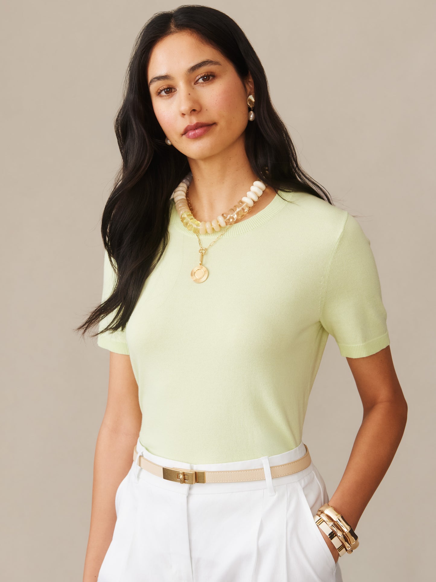 A woman wearing the Joelle Sweater by J.McLaughlin in a soft celery green shade, styled with white pants and gold accessories, showcasing effortlessly stylish, enduring designs with innovative fabrics, a destination for defining style, life well-lived.