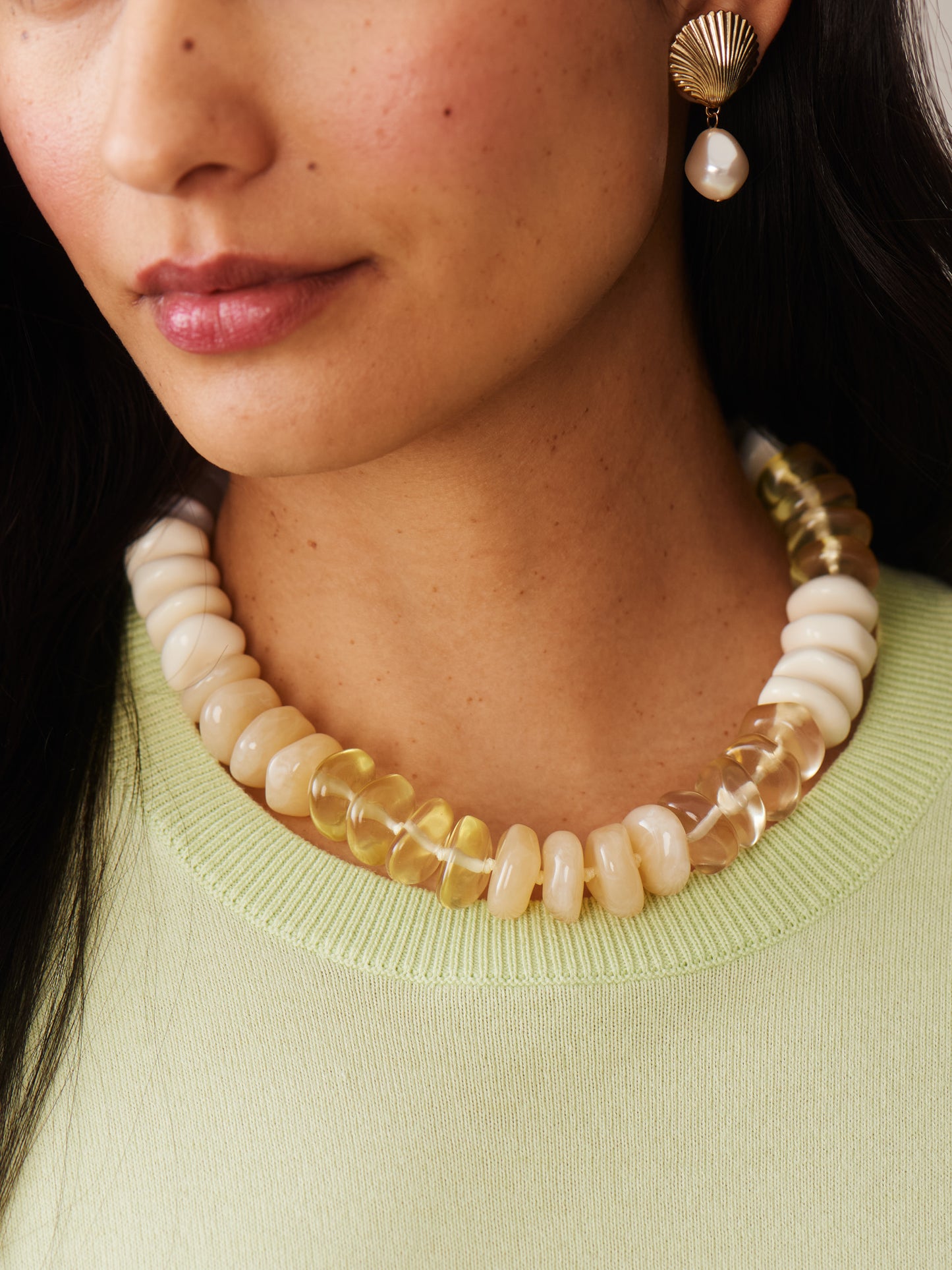 Close-up of a woman wearing a Tonal Bead Necklace by J.McLaughlin featuring smooth round beads in soft cream and translucent amber hues, paired with a light green top and a gold and pearl earring.